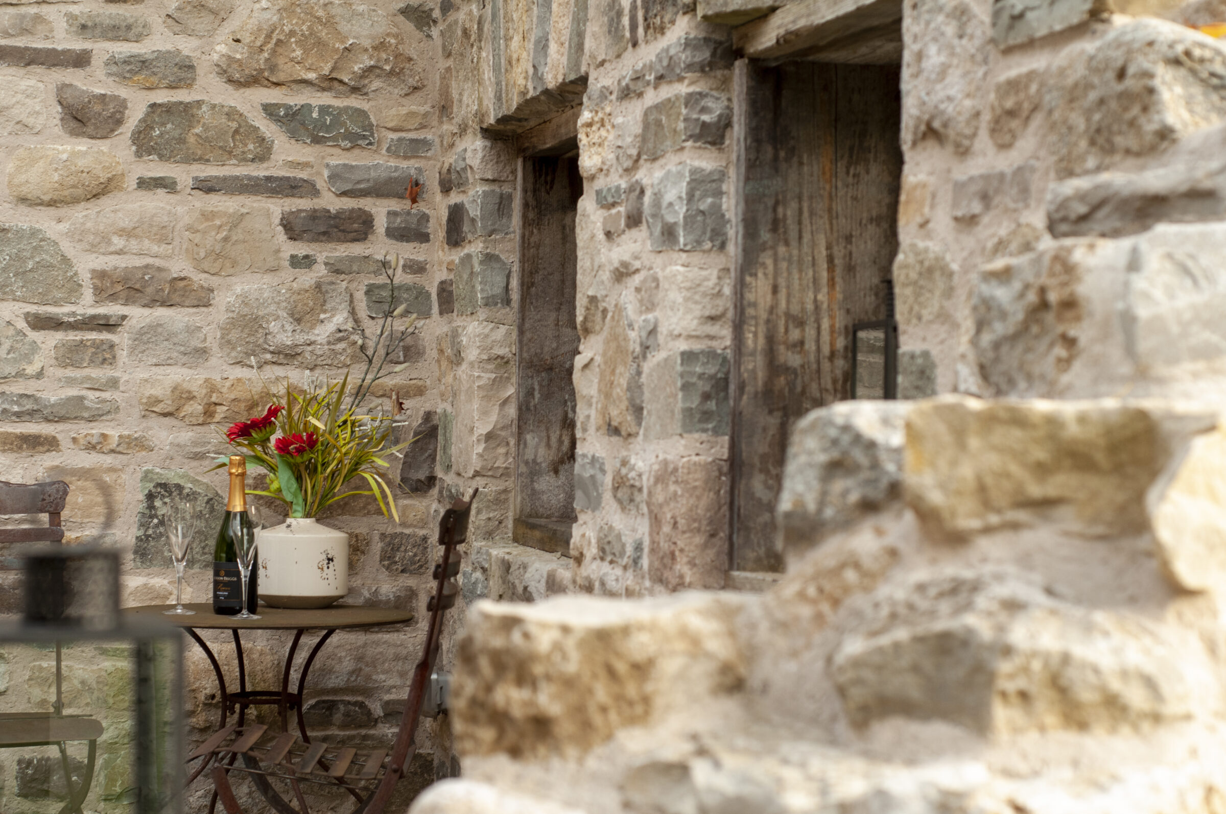 A rustic stone patio features a small table, two chairs, a champagne bottle, and a vase with red flowers.
