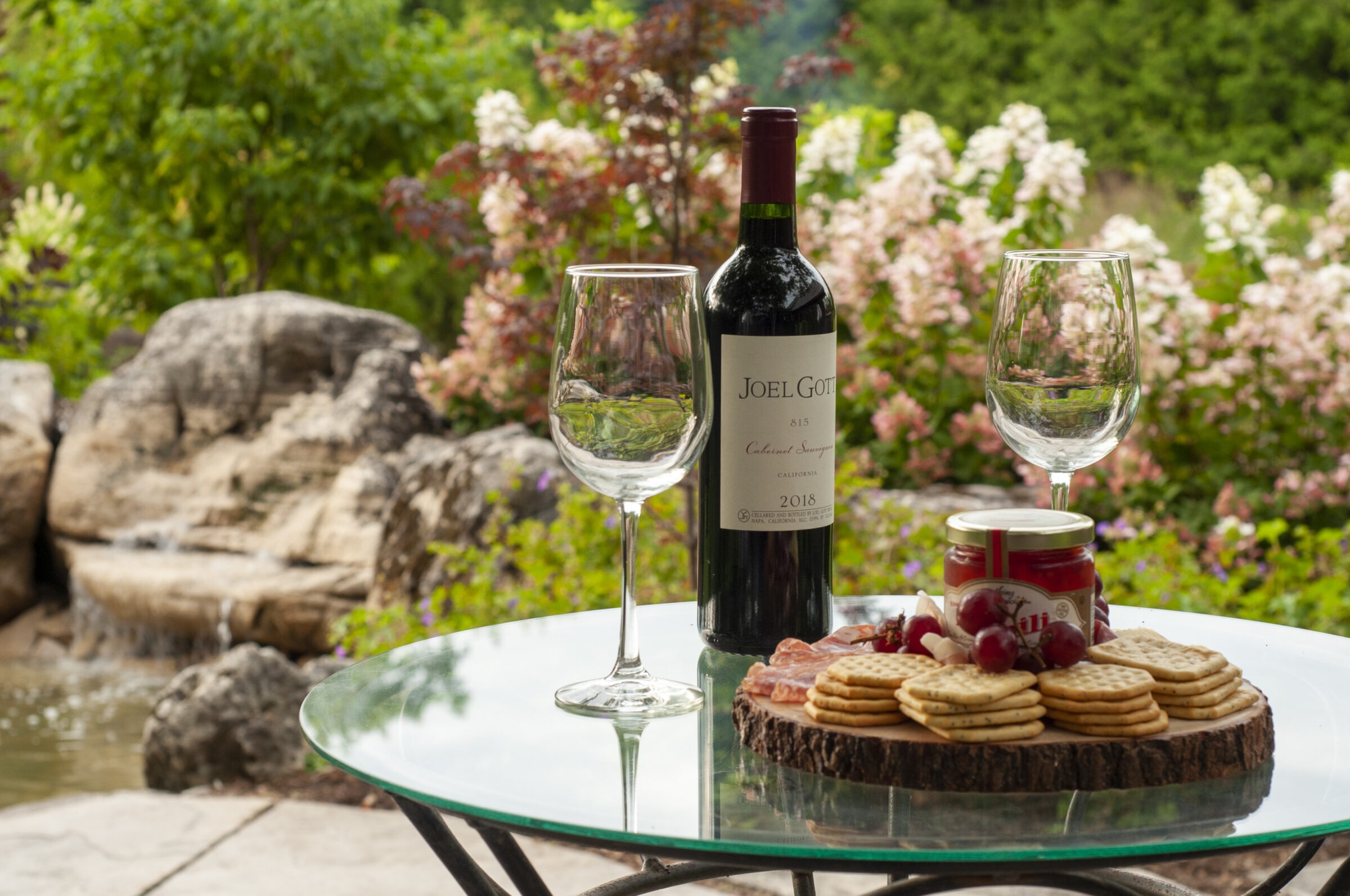 A rustic outdoor setting features wine, two glasses, and a wooden platter of crackers and grapes on a glass table, surrounded by flowers.
