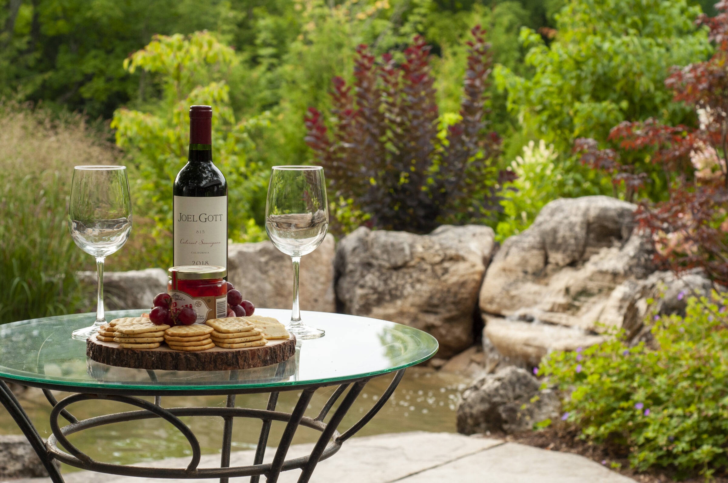 A glass table with wine, two glasses, crackers, and grapes, set outdoors with lush greenery and rocks in the background.