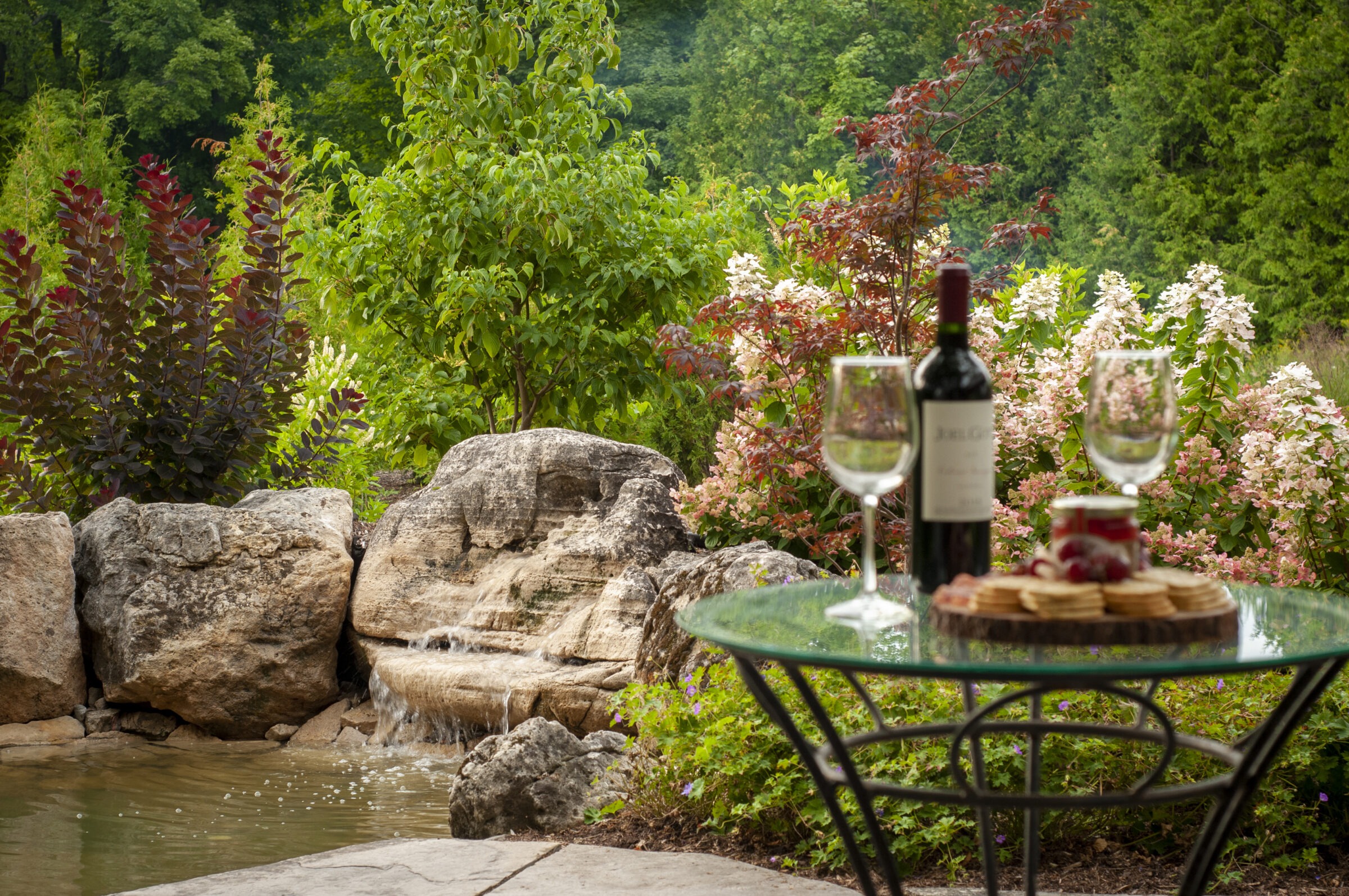 A serene garden with water feature, surrounded by lush greenery. Glass table holds wine, glasses, and snacks, offering a tranquil setting.