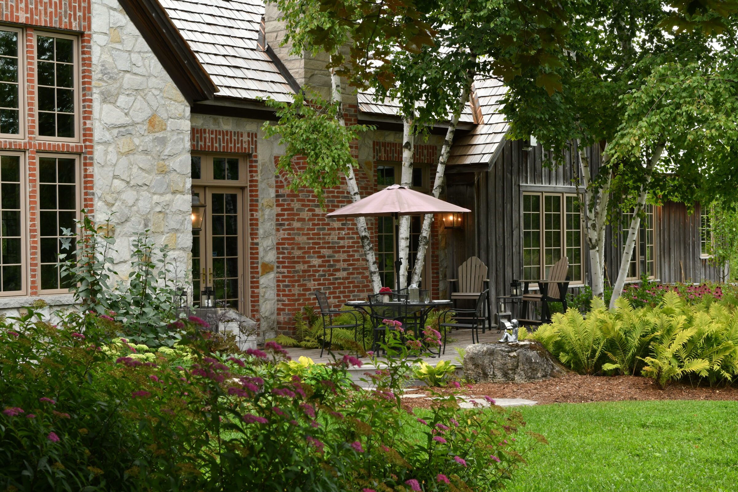 Charming house with stone and brick walls, surrounded by lush greenery and flowers. Patio features outdoor seating with umbrella, inviting atmosphere.