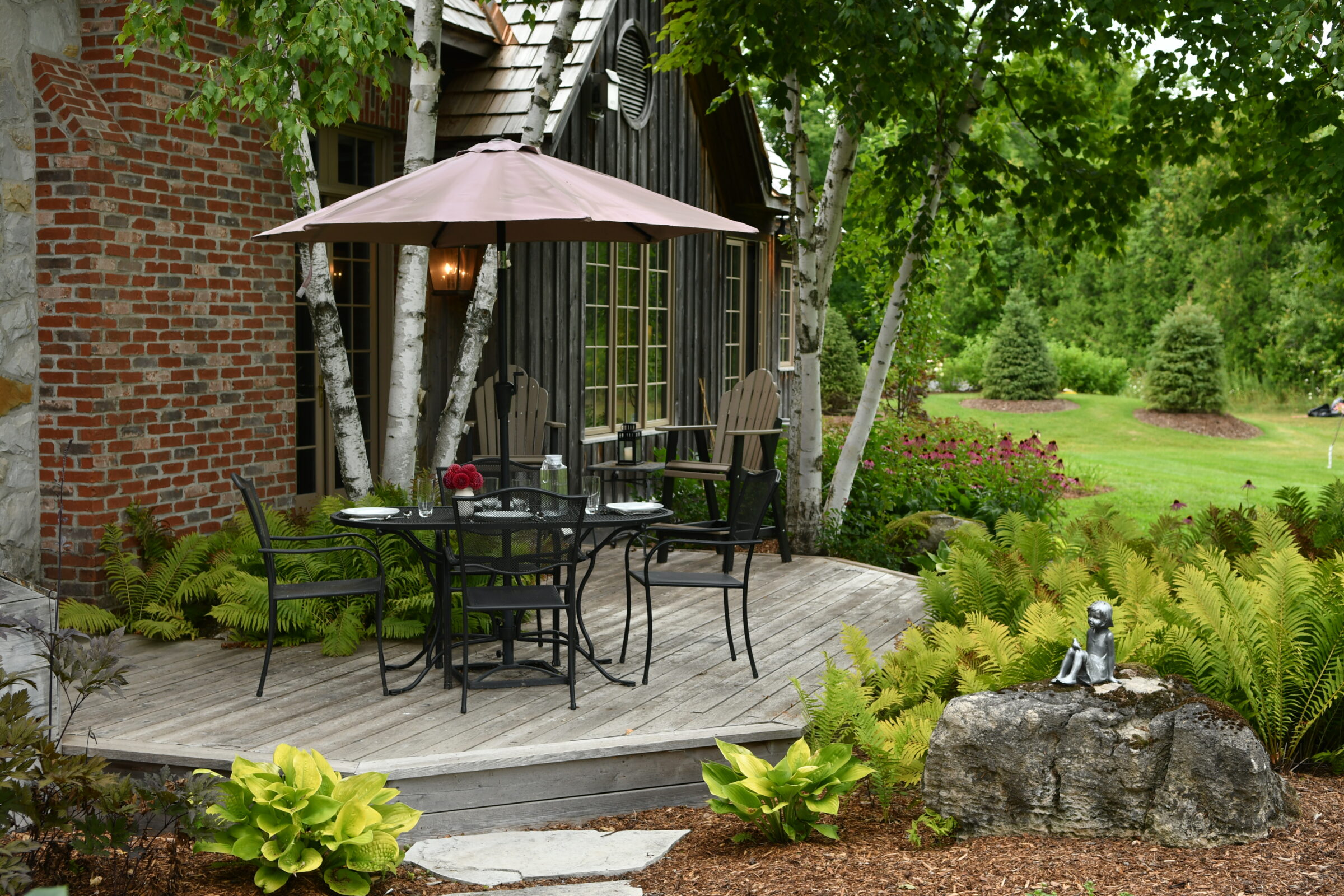 A peaceful garden scene with patio furniture, an umbrella, lush greenery, and a small sculpture on a rock beside a house.