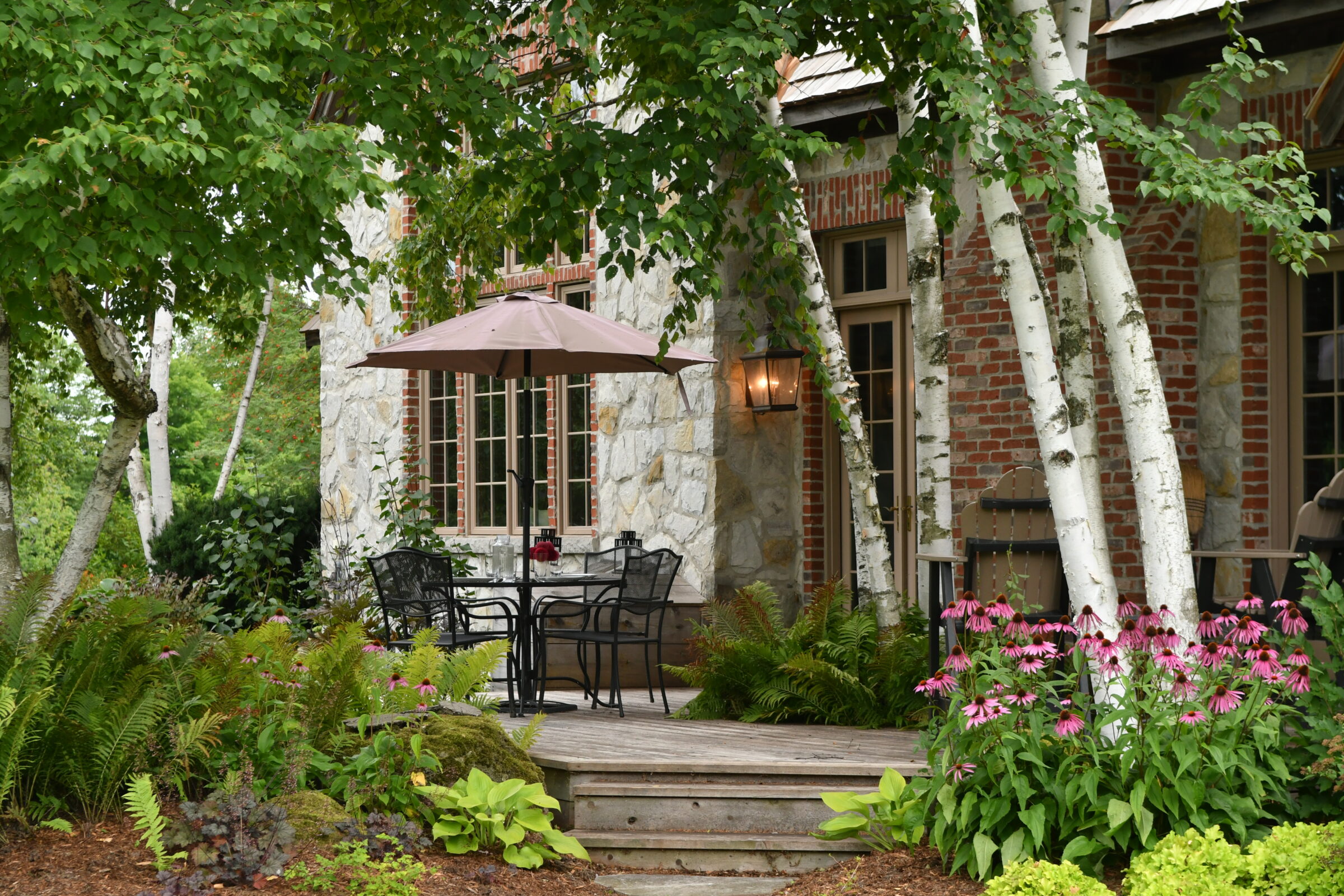 A peaceful garden with a stone house, wooden deck, patio furniture, umbrella, birch trees, and vibrant flowers under green foliage.