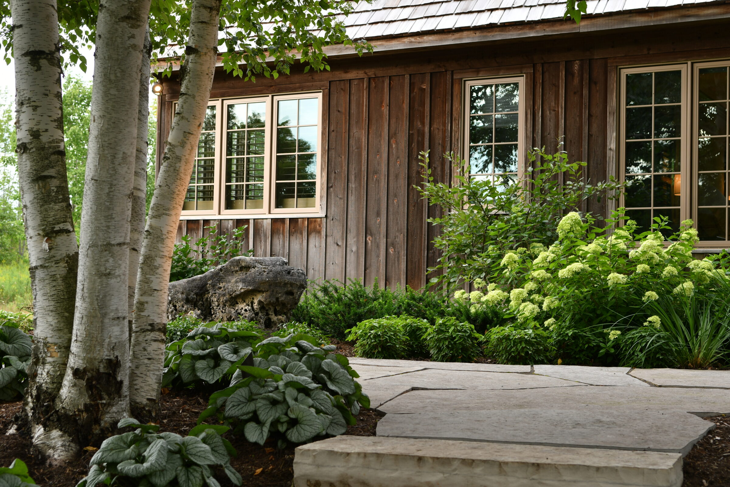 Rustic wooden house with large windows surrounded by lush greenery, birch trees, and a stone path in a serene garden setting.