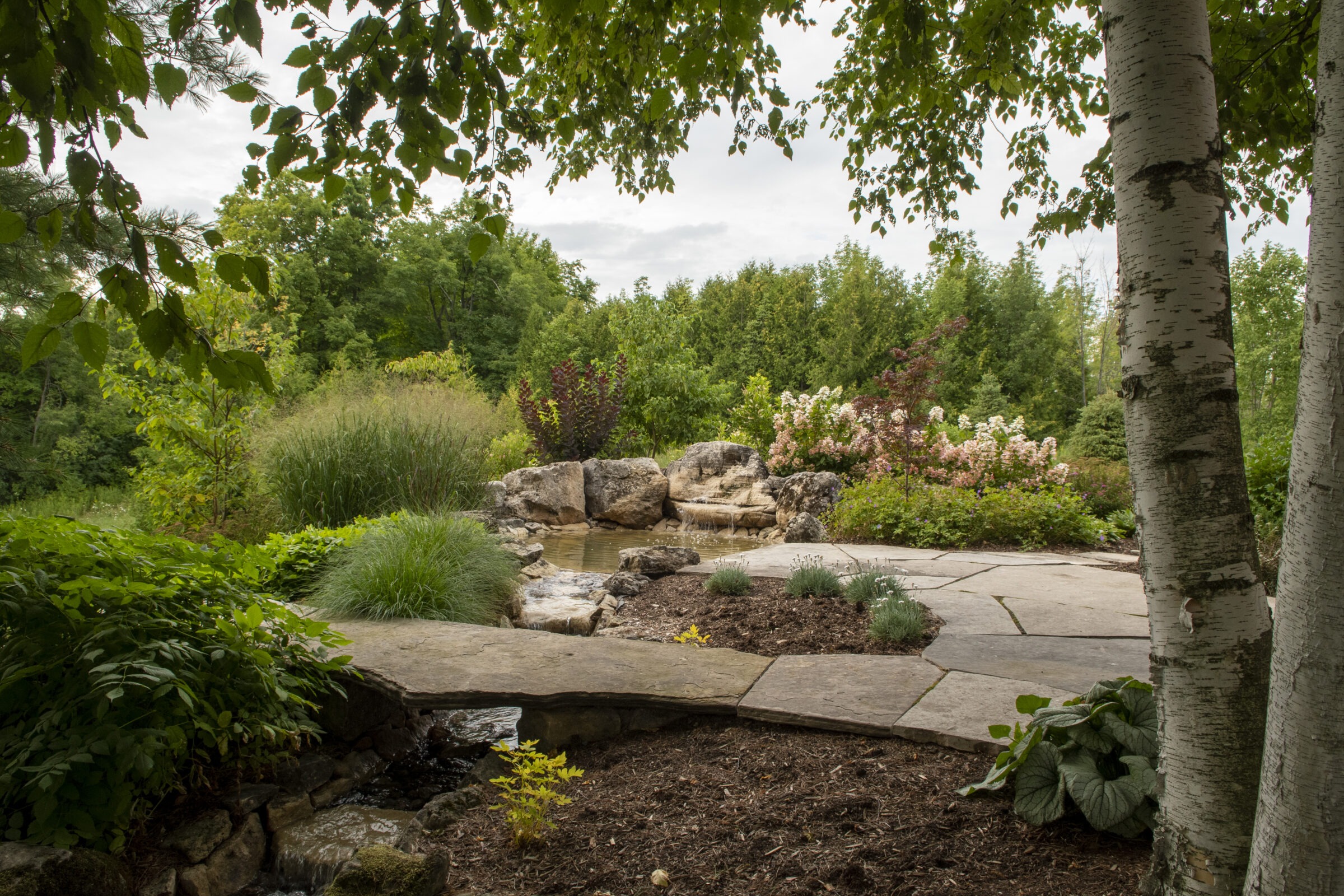 A tranquil garden scene with a stone path, flowing water feature, lush greenery, and colorful blooms beneath a partially cloudy sky.