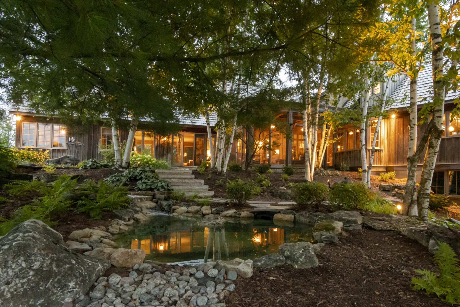 A cozy wooden house surrounded by trees and a small pond, illuminated warmly in the evening, creating a serene natural setting.