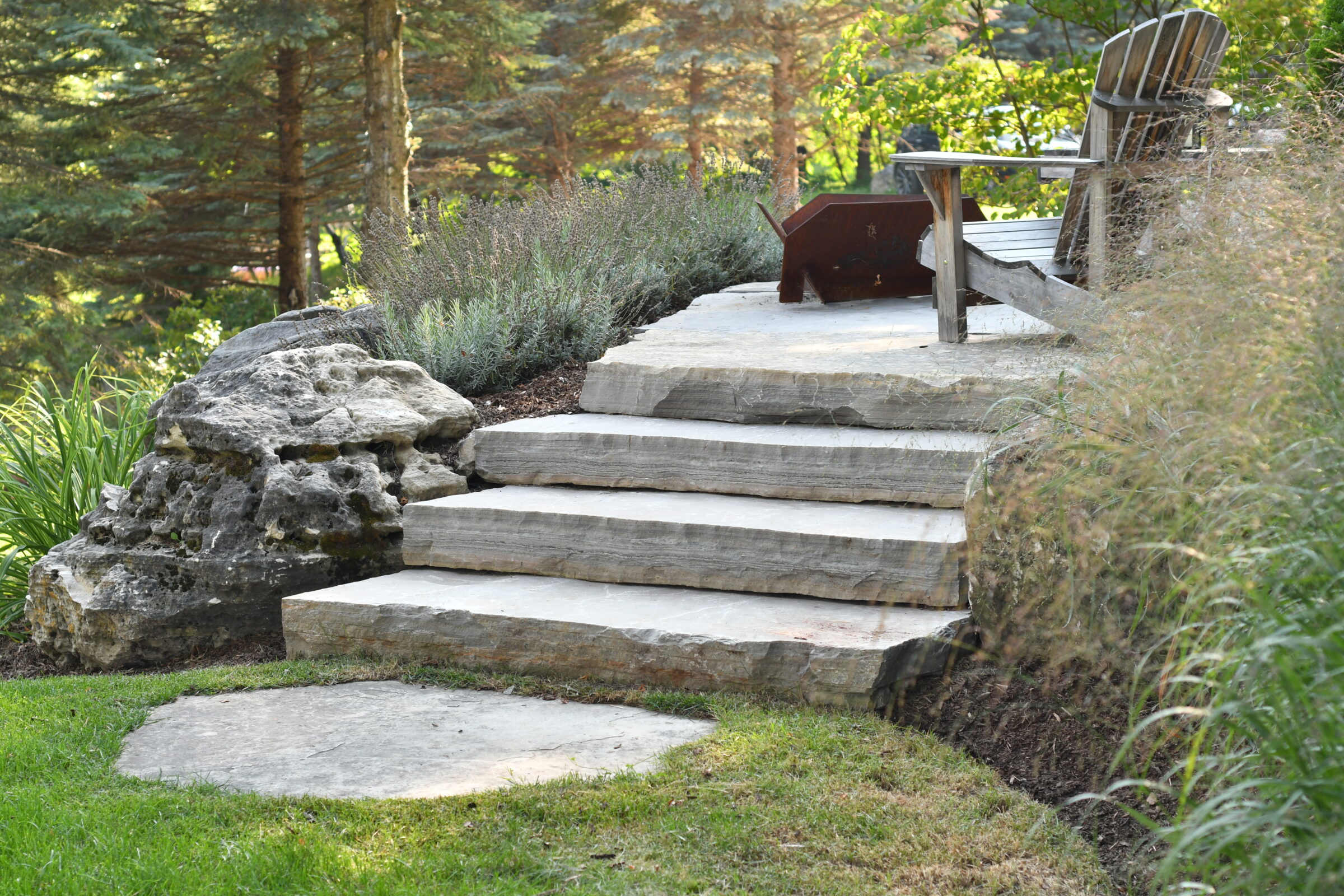 Stone steps lead to rustic wooden chairs surrounded by greenery and trees, creating a serene natural outdoor setting.