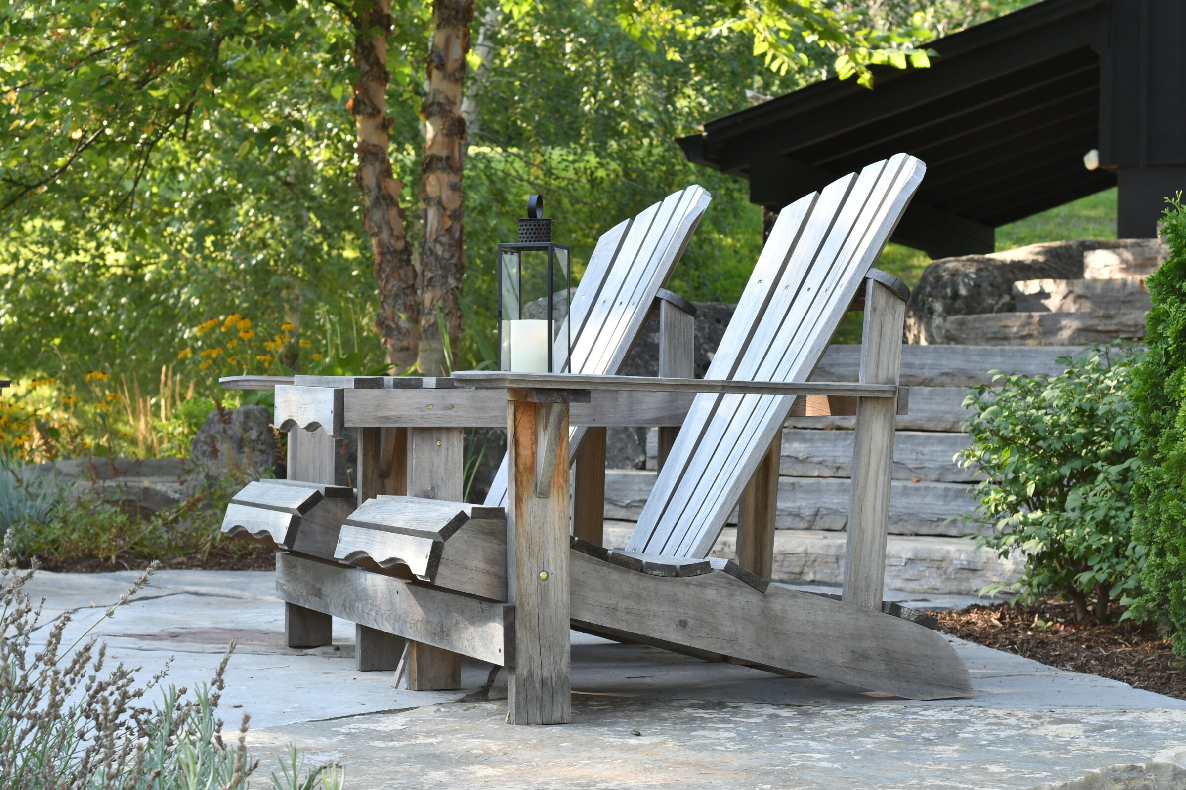 Rustic wooden Adirondack chairs with a lantern on stone patio, surrounded by lush greenery and trees, under a clear sky. Serene outdoor setting.