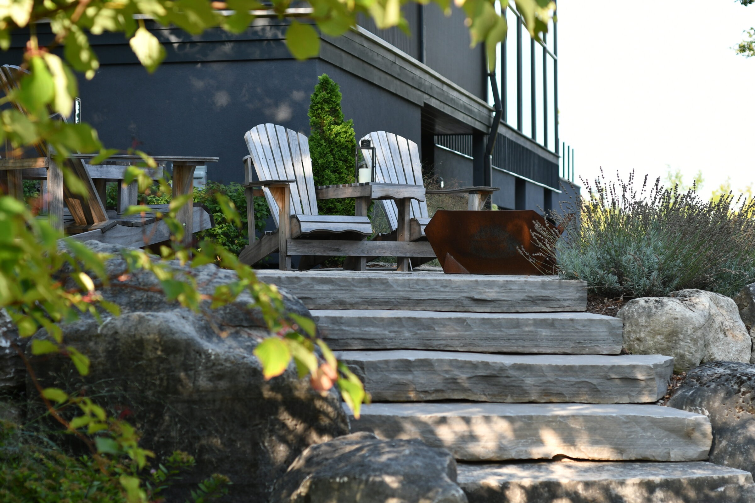 Two wooden Adirondack chairs on stone steps, surrounded by lush greenery and rocks, outside a modern building with large windows.