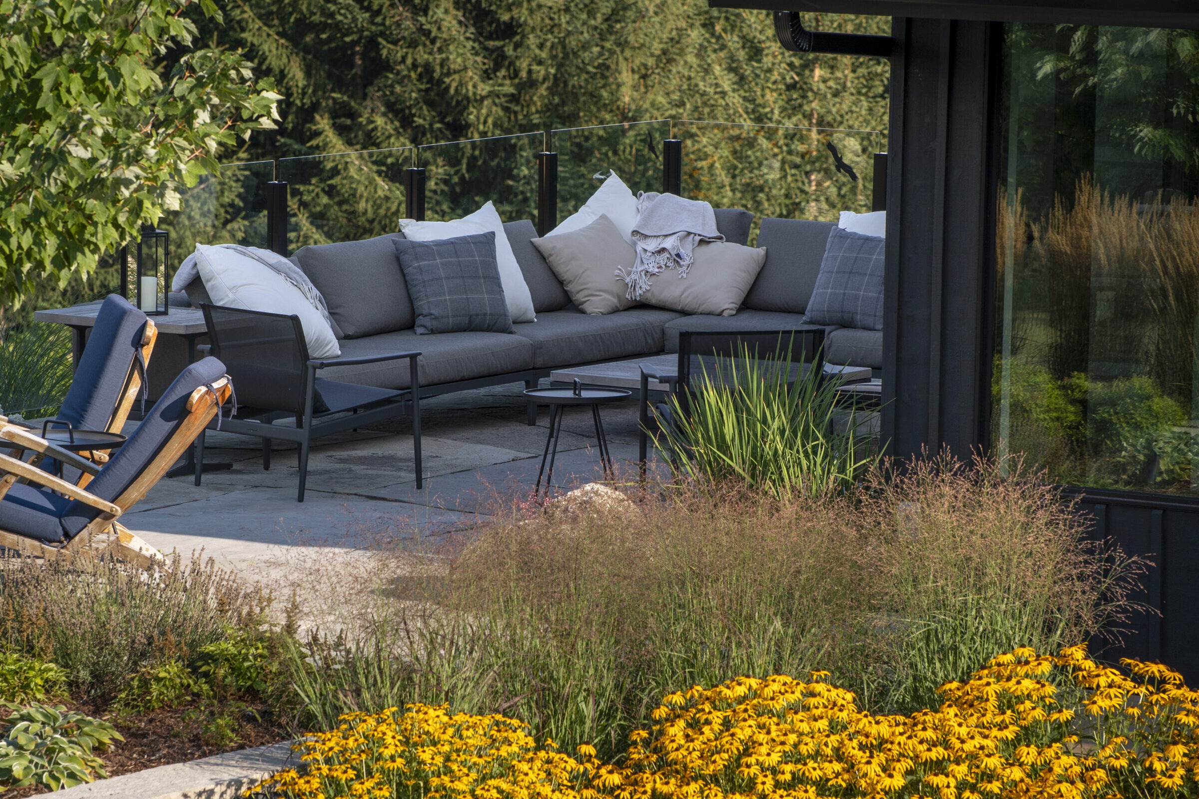 A cozy outdoor patio with cushioned seating, surrounded by lush greenery and yellow flowers, offering a relaxing view of the forest.