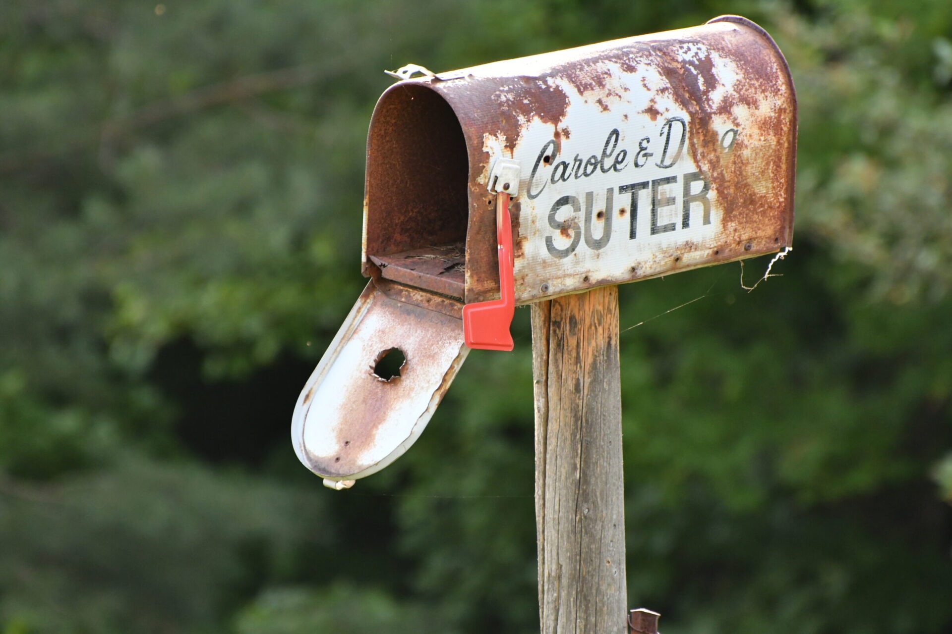 Rusty mailbox on a wooden post against greenery. Red flag is raised. The name 