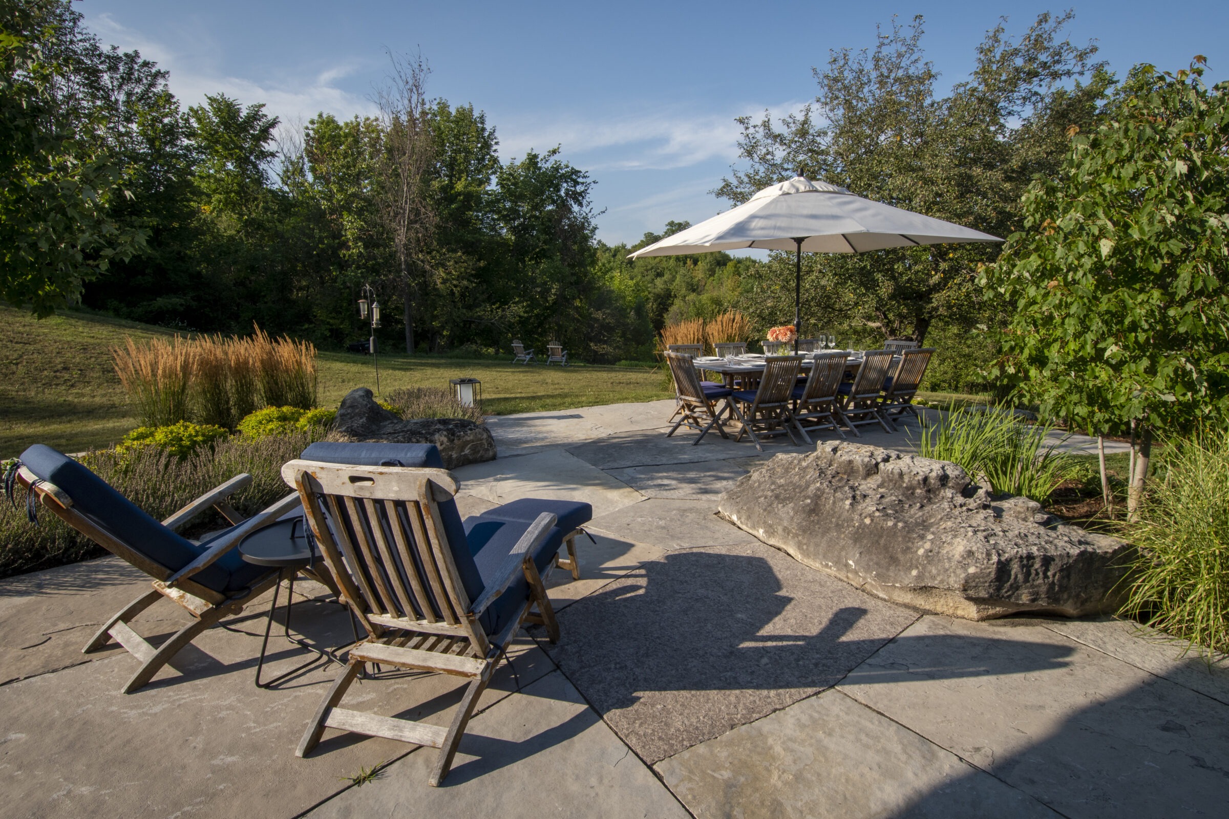 Patio scene with lounge chairs and a dining table under an umbrella, set in a lush garden with trees and greenery.
