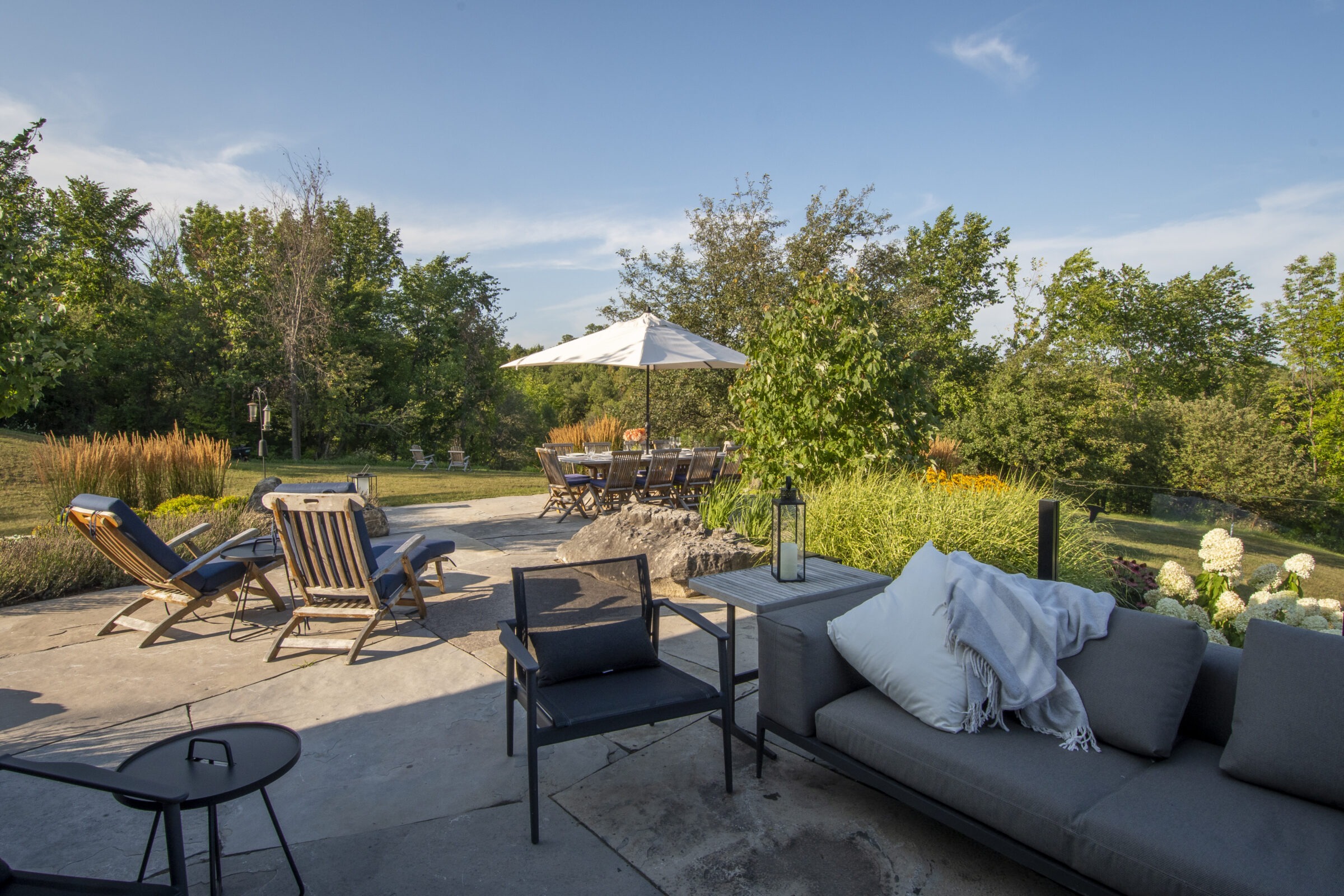 A tranquil patio scene with loungers, a dining area under an umbrella, lush greenery, and comfortable seating, set in a sunny garden.