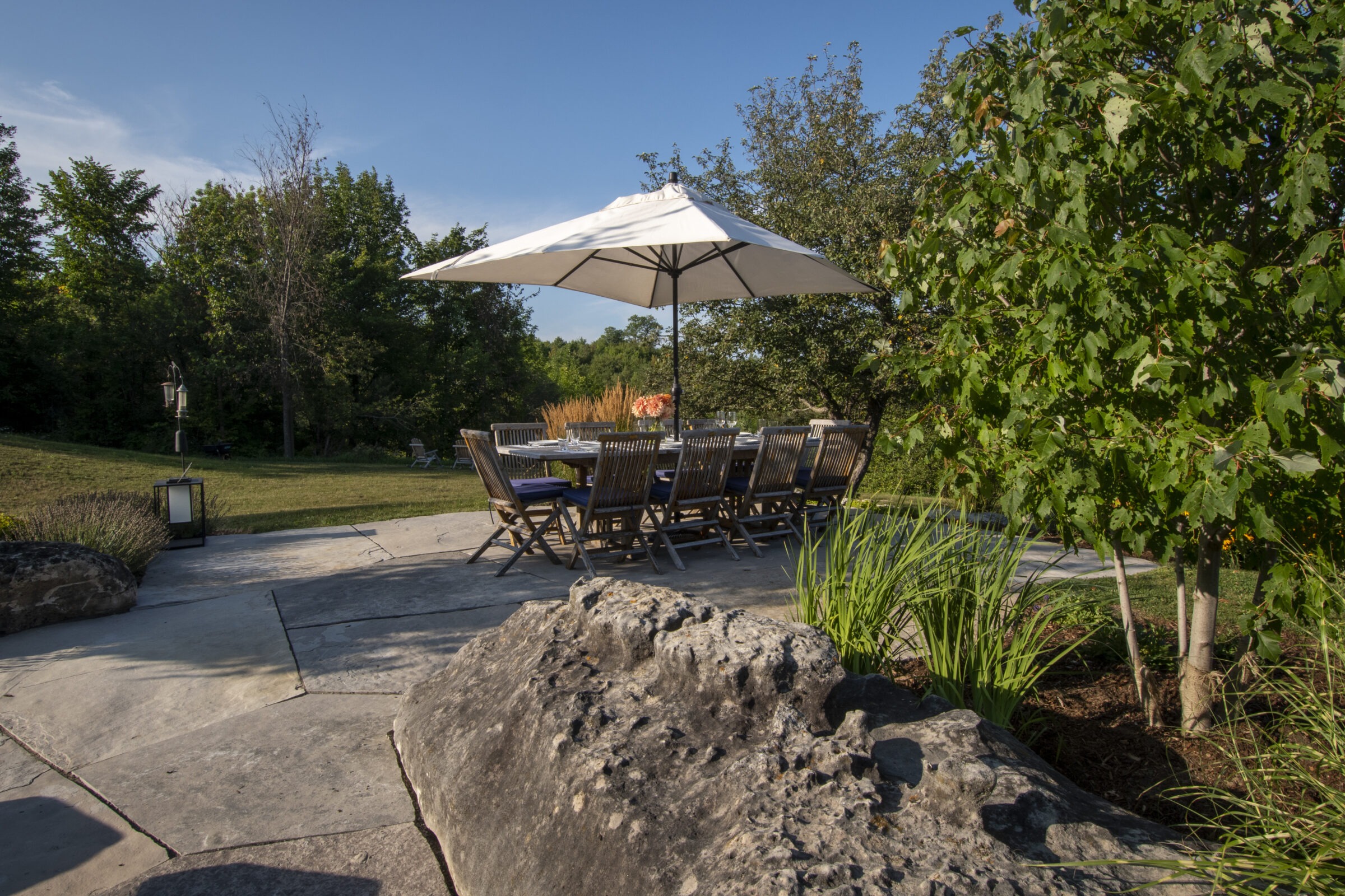 Outdoor dining setup with wooden chairs and umbrella on stone patio, surrounded by lush greenery and trees under a clear blue sky.