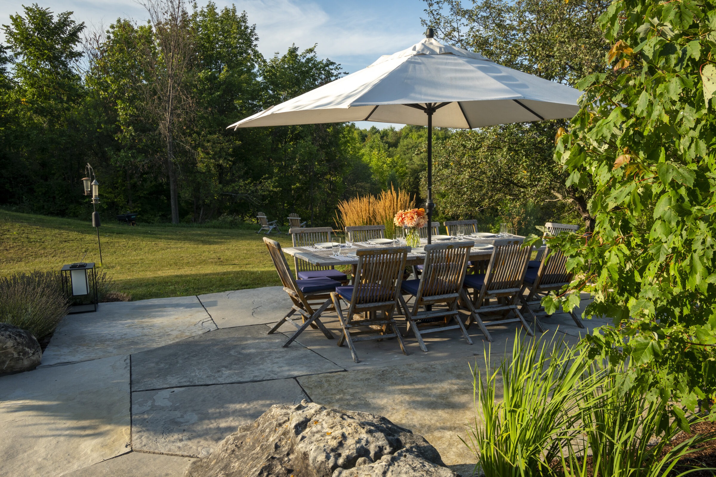 A patio with a large table and umbrella, surrounded by greenery and trees, set for dining outdoors in a natural setting.