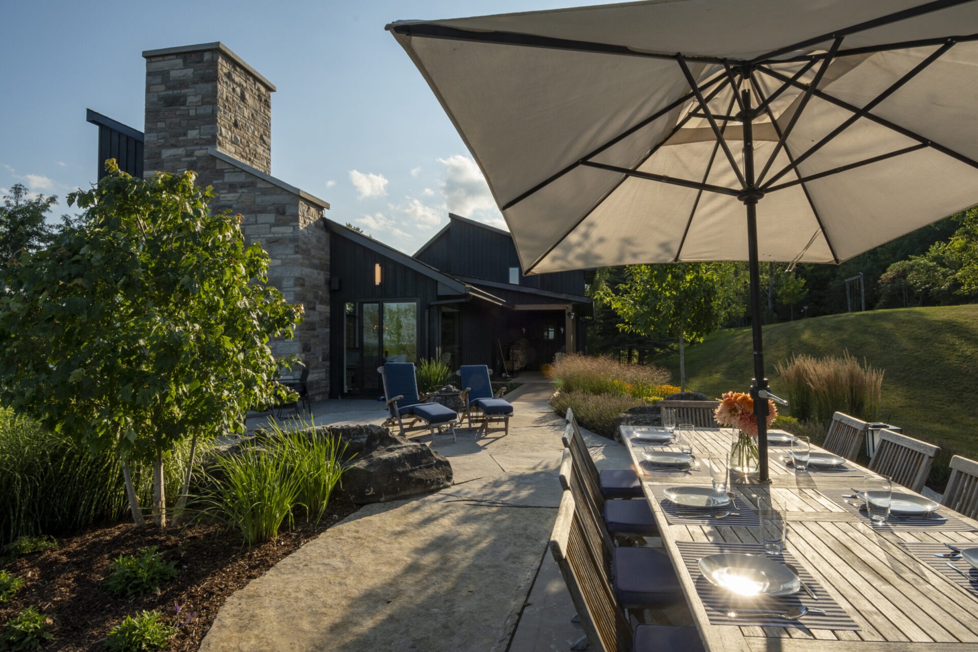 A modern patio with a long dining table, umbrella, stone fireplace, and greenery. Sunlit and peaceful outdoor space perfect for gatherings.