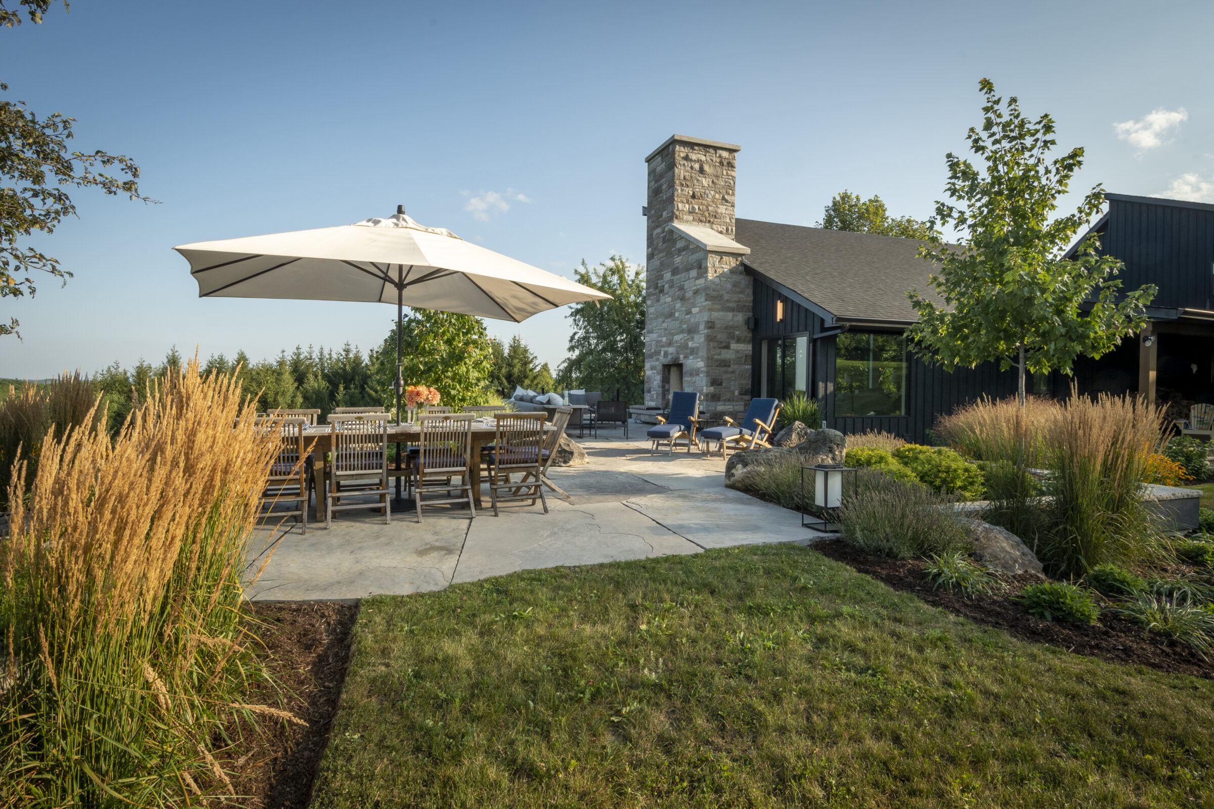 Spacious patio with outdoor dining, umbrella, and seating. Stone chimney, greenery, and wooden accents create a relaxing garden atmosphere under a clear blue sky.