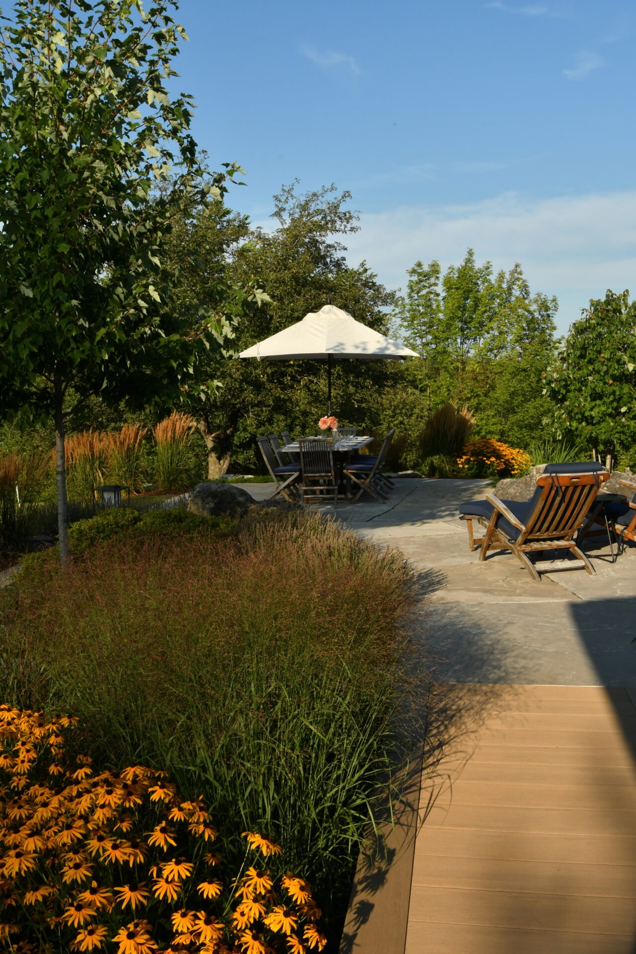 Patio scene with table, chairs, and umbrella. Surrounded by lush greenery and bright flowers on a sunny day, providing a peaceful outdoor setting.