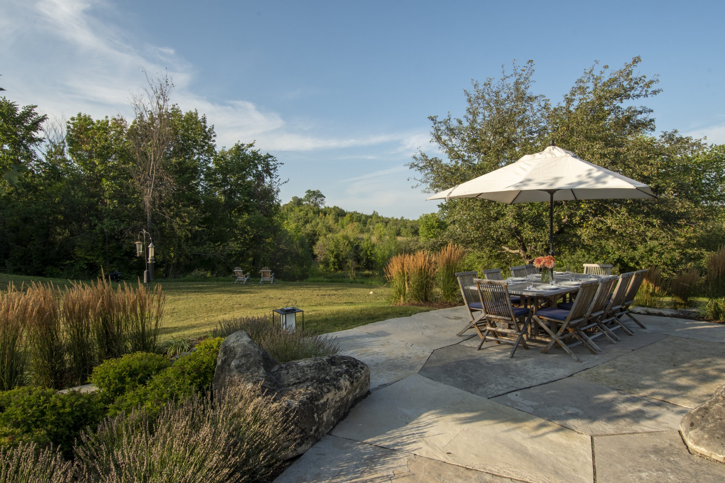 A tranquil backyard patio with a large table, chairs, umbrella, and surrounding lush greenery, under a bright blue sky.