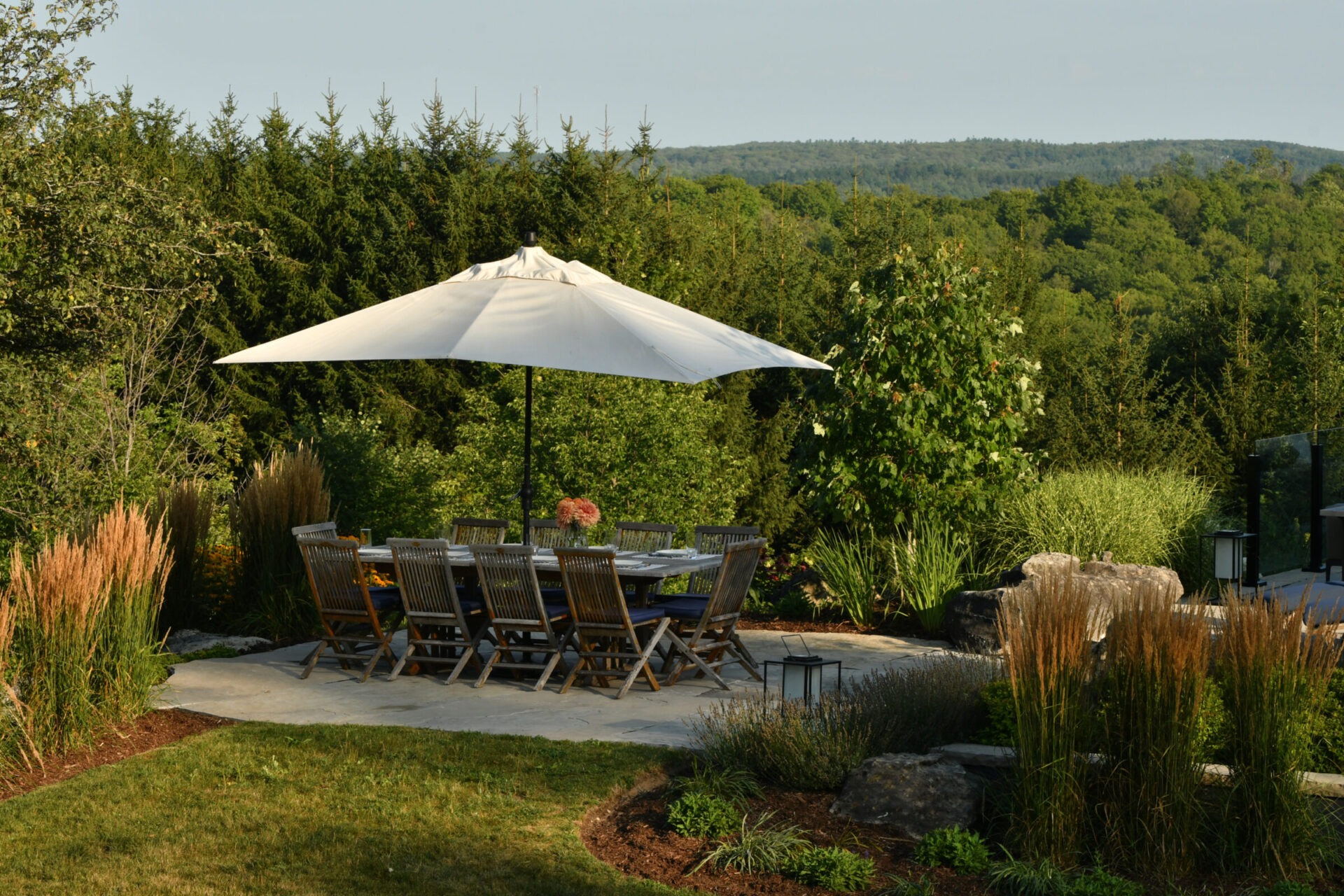 A serene outdoor dining area with a large umbrella and wooden chairs, surrounded by lush greenery and distant rolling hills.
