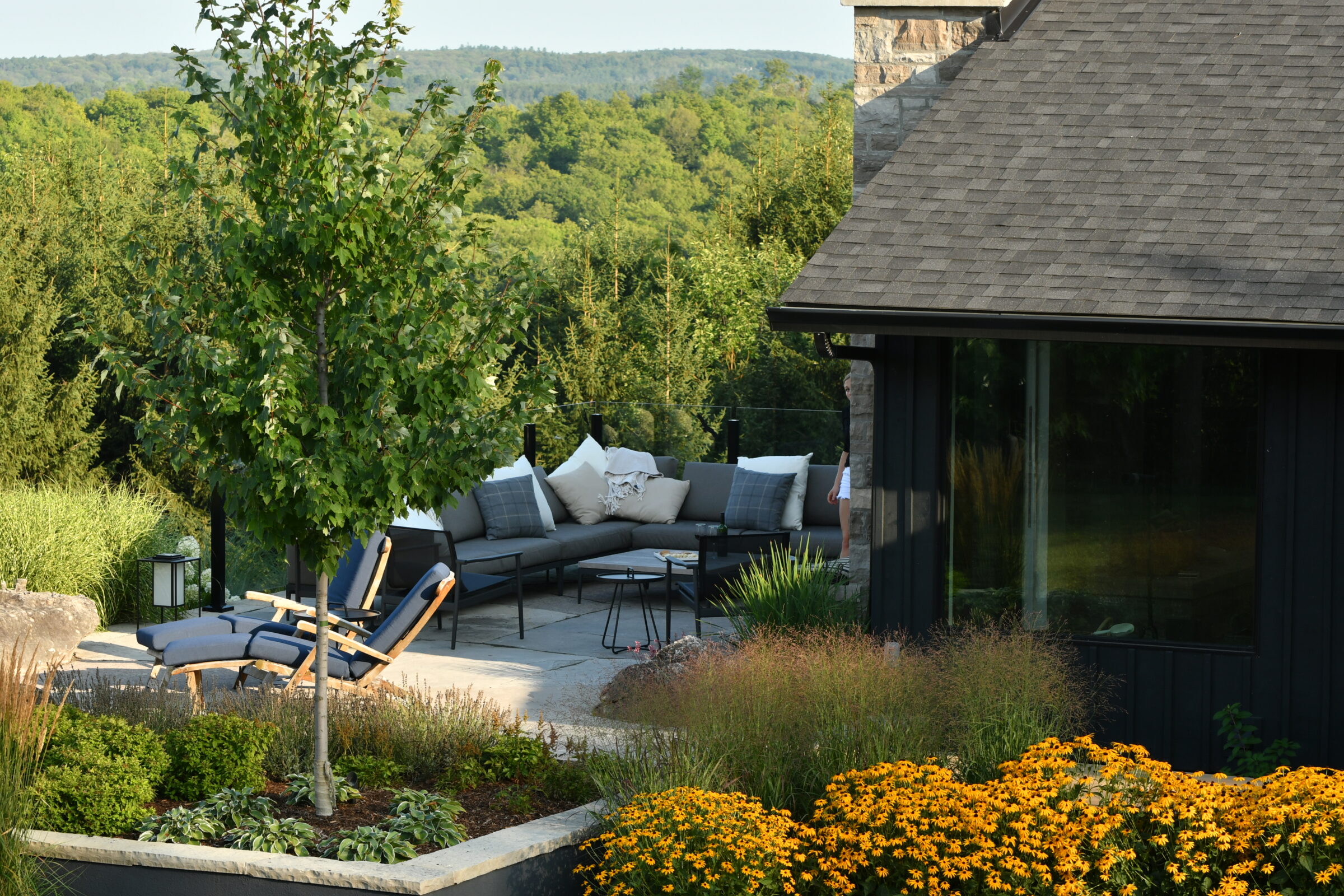 Terrace with lounge seating and colorful flowers adjacent to a modern house. Scenic forested hills in the background under a clear sky.