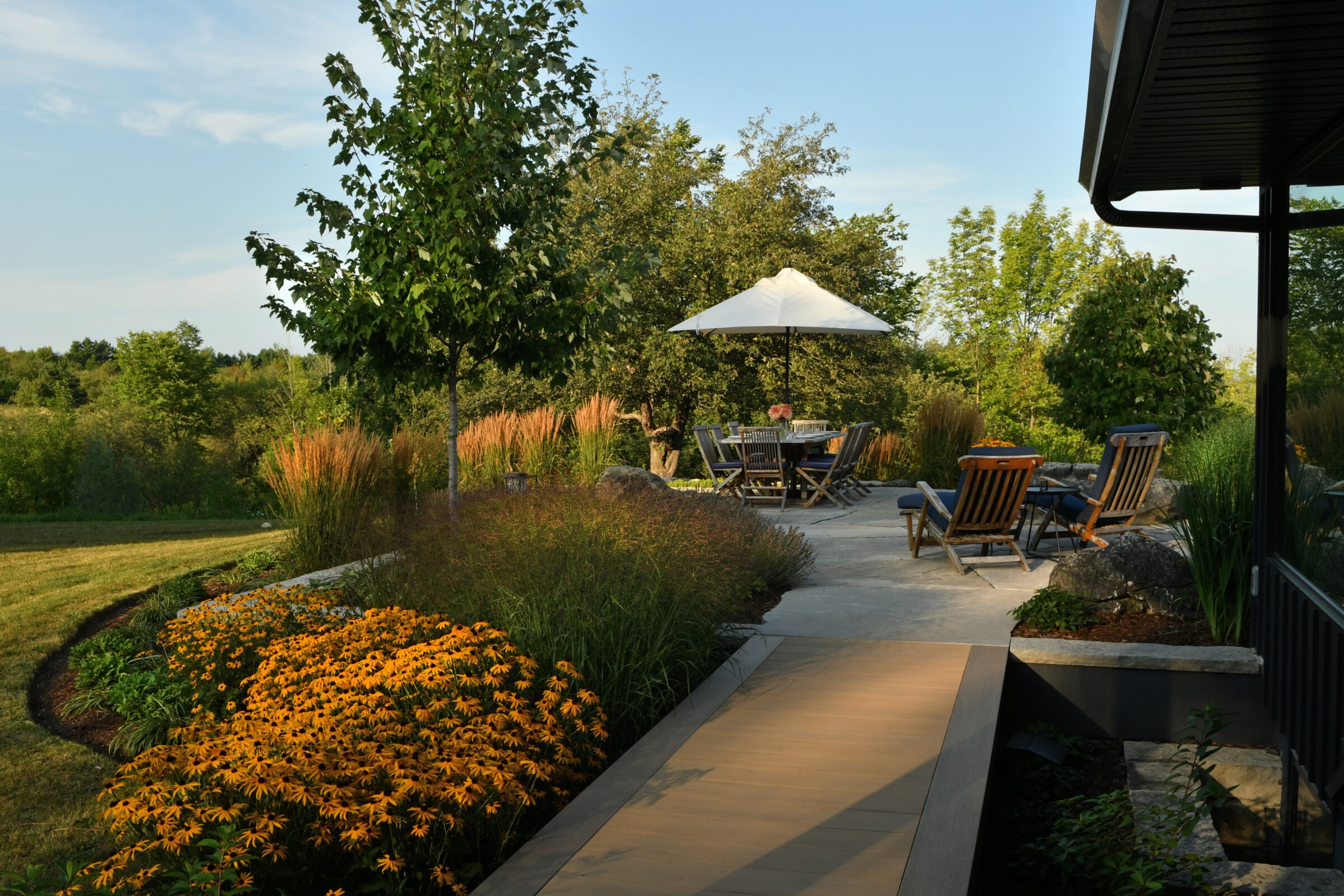 A peaceful garden scene with patio furniture, colorful flowers, and lush greenery on a sunny day, featuring a shaded seating area.