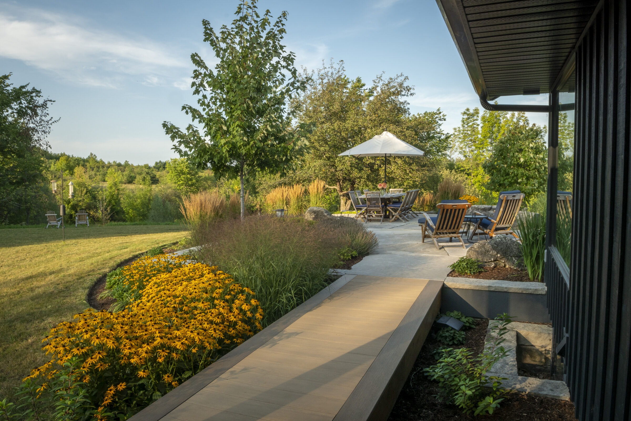 A serene garden scene with yellow flowers, a wooden walkway, outdoor seating under an umbrella, and lush greenery under a clear sky.