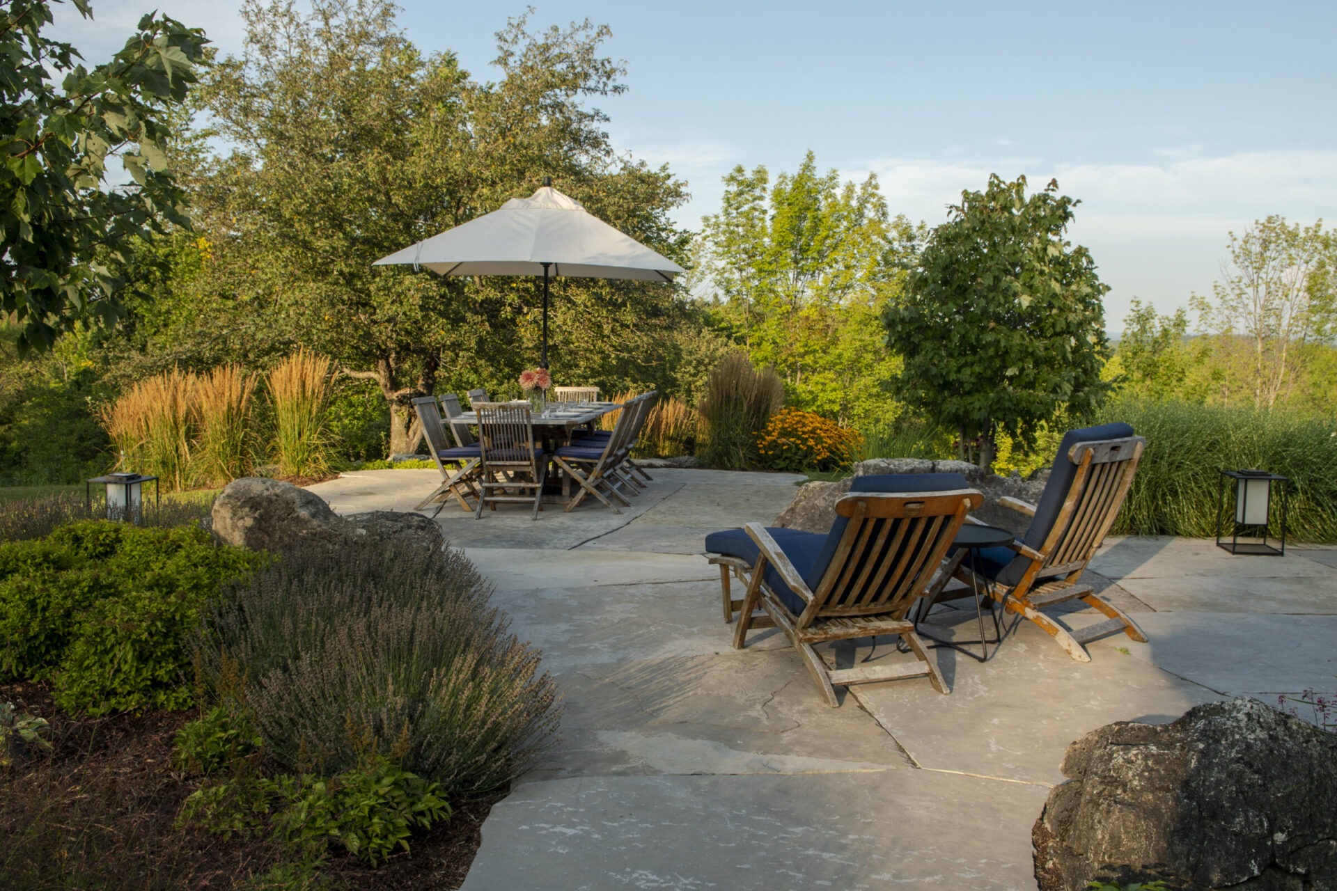 A serene patio with wooden chairs, table, and umbrella amidst lush greenery and trees on a sunny day.