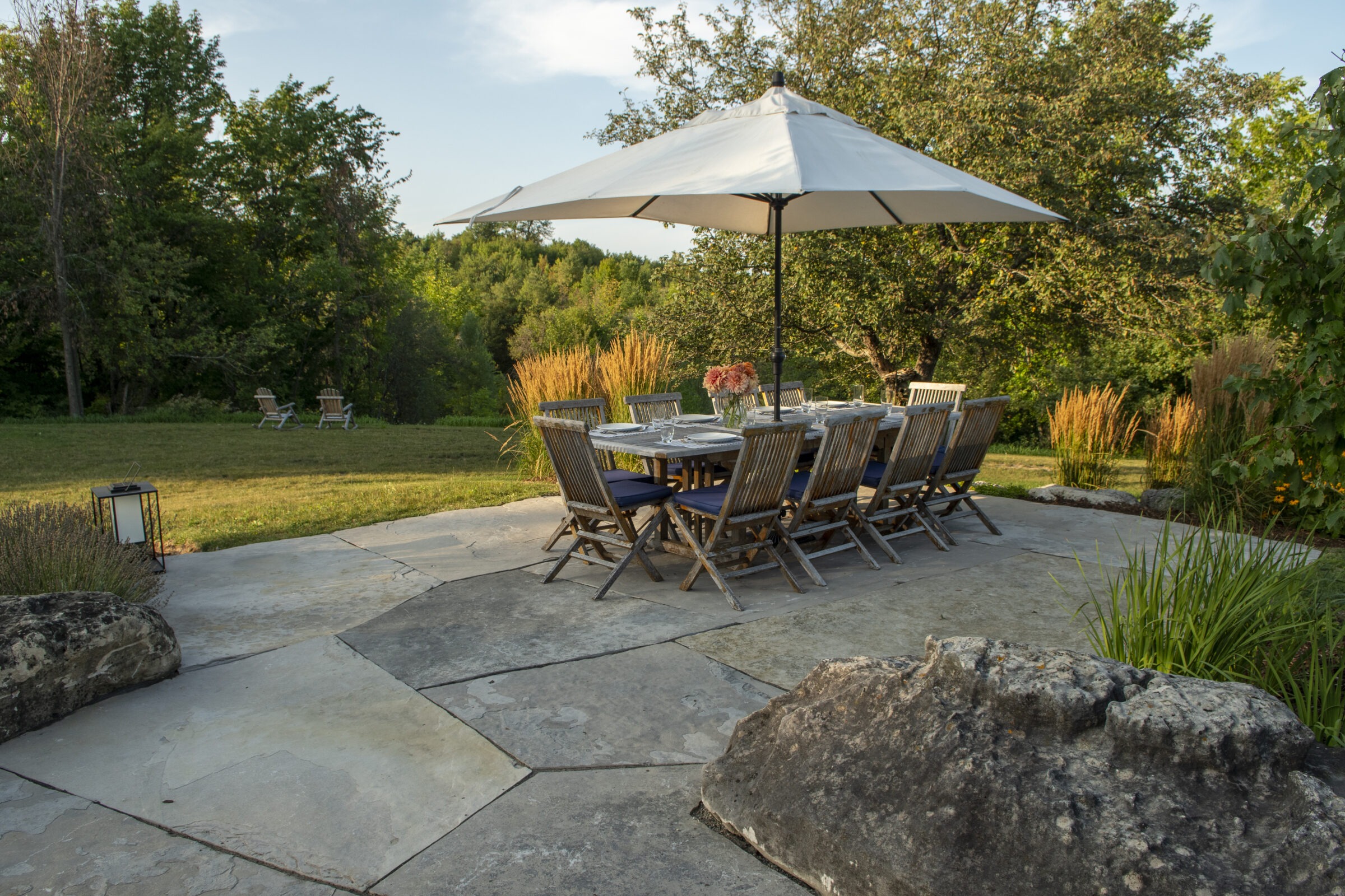 A patio set with wooden chairs and table under an umbrella, surrounded by lush greenery and boulders, with a peaceful outdoor setting.