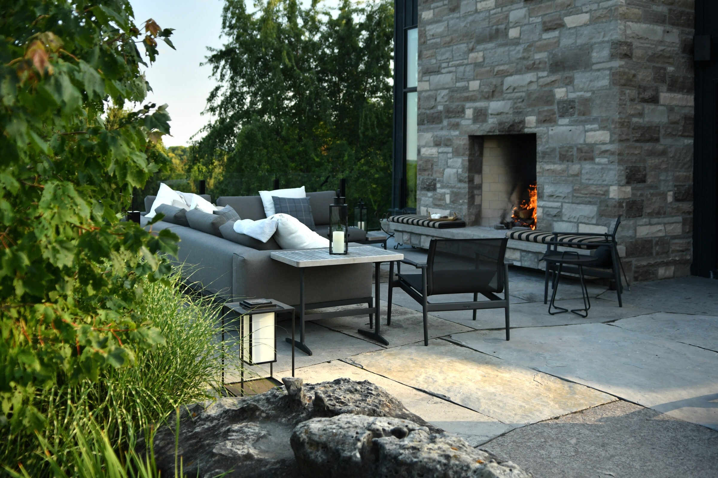 Modern outdoor patio with stone fireplace, cozy seating, and lush greenery. Sofa with cushions, table with lantern, and vibrant plants create a serene atmosphere.