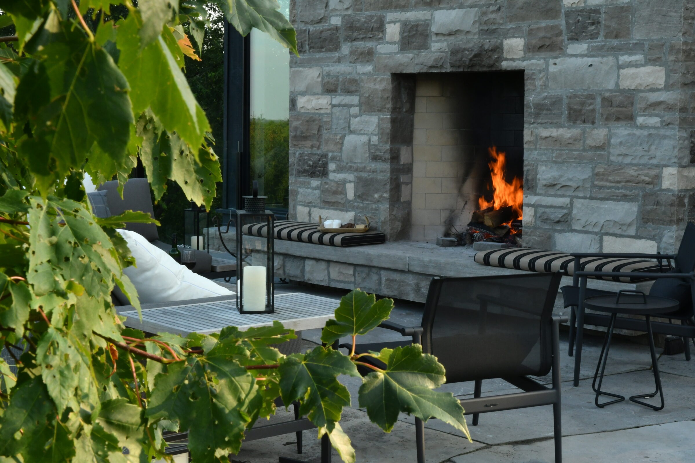 Outdoor patio with modern furniture, a stone fireplace, and a small fire. Green leaves partially frame the cozy seating area.