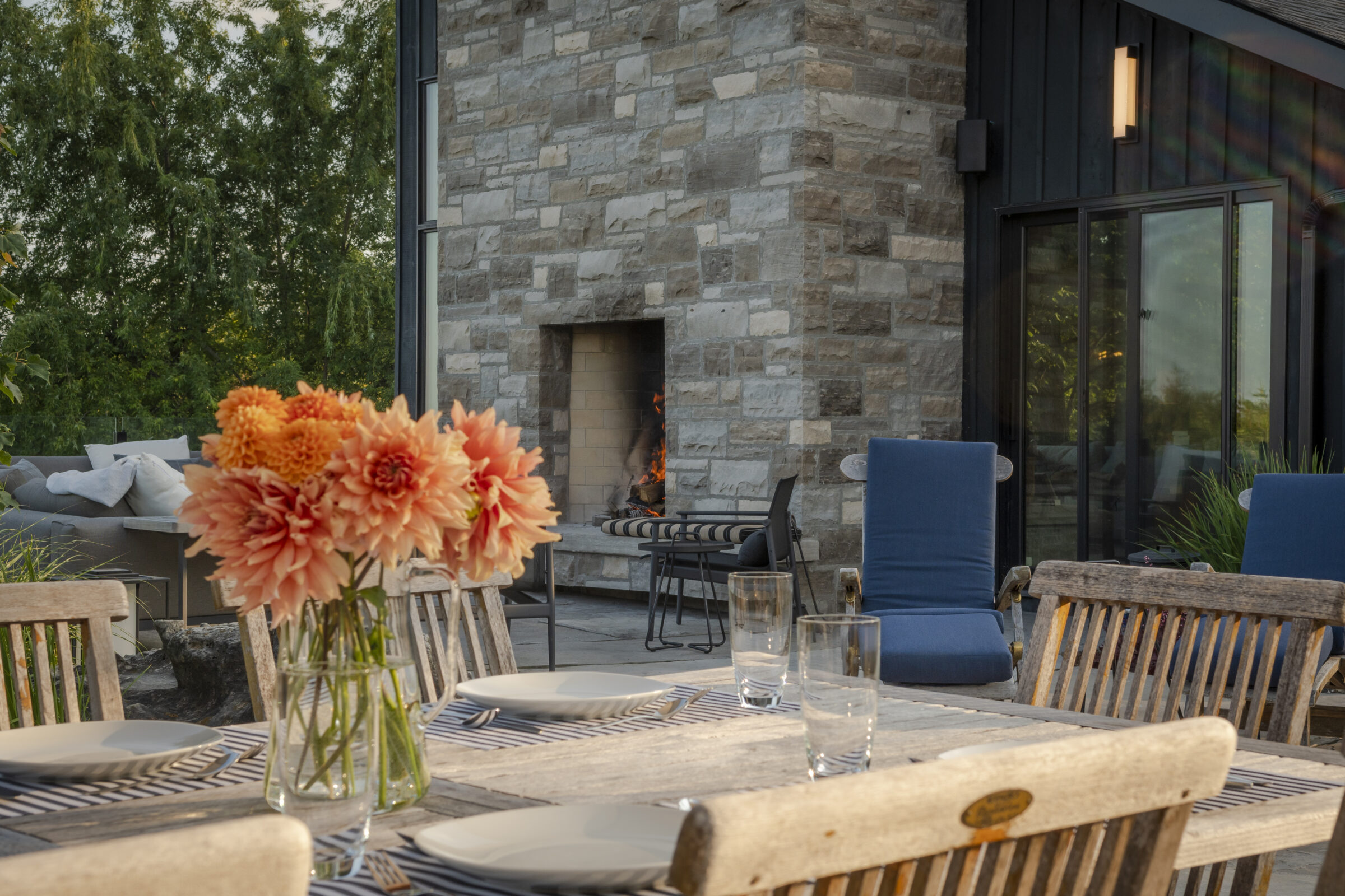 A cozy outdoor patio with a stone fireplace, wooden dining table, striped cushions, and vase of flowers, surrounded by lush green trees.