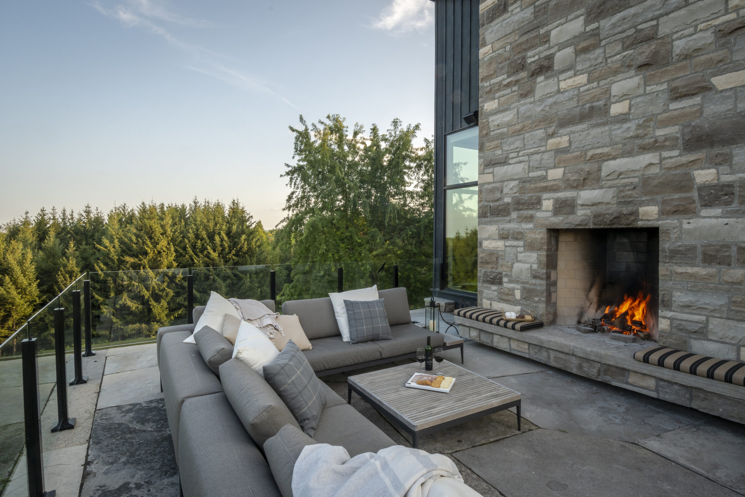 Outdoor patio with a stone fireplace, comfortable seating, and scenic forest view. Wine and cheese on the table, under a clear sky.