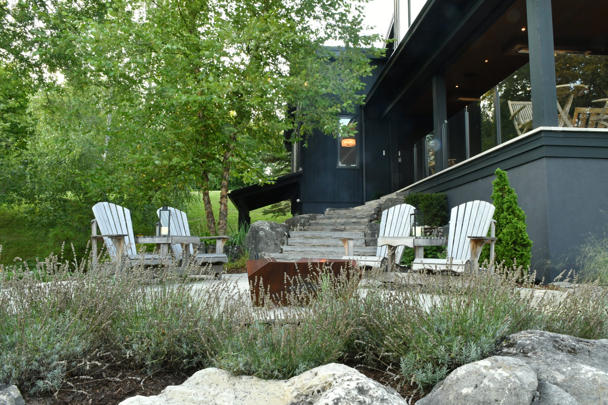 Modern house patio with Adirondack chairs, fire pit, and lush greenery. Stone steps lead to an outdoor living area, creating a tranquil setting.