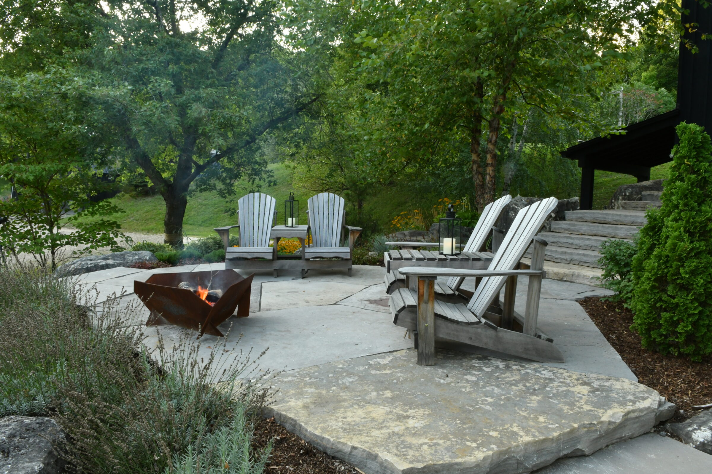 A serene outdoor patio with wooden chairs, a fire pit, and lush greenery, perfect for relaxation amidst natural surroundings.