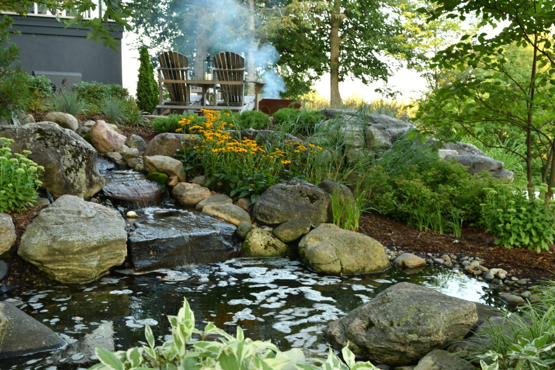Tranquil garden scene with a small waterfall, vibrant flowers, and two Adirondack chairs beside a smoking fire pit under lush green trees.