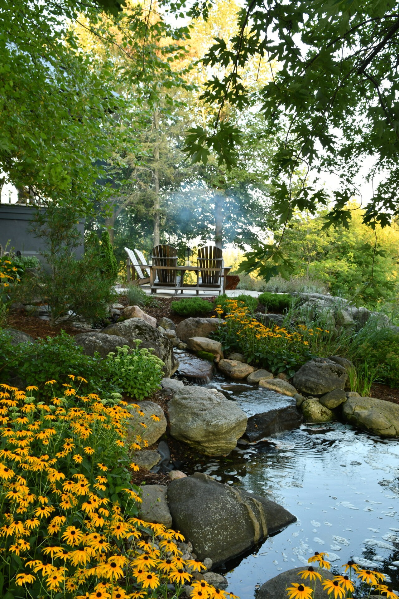 Tranquil garden with Adirondack chairs, vibrant yellow flowers, rocks, and a small stream, surrounded by lush green trees on a sunny day.