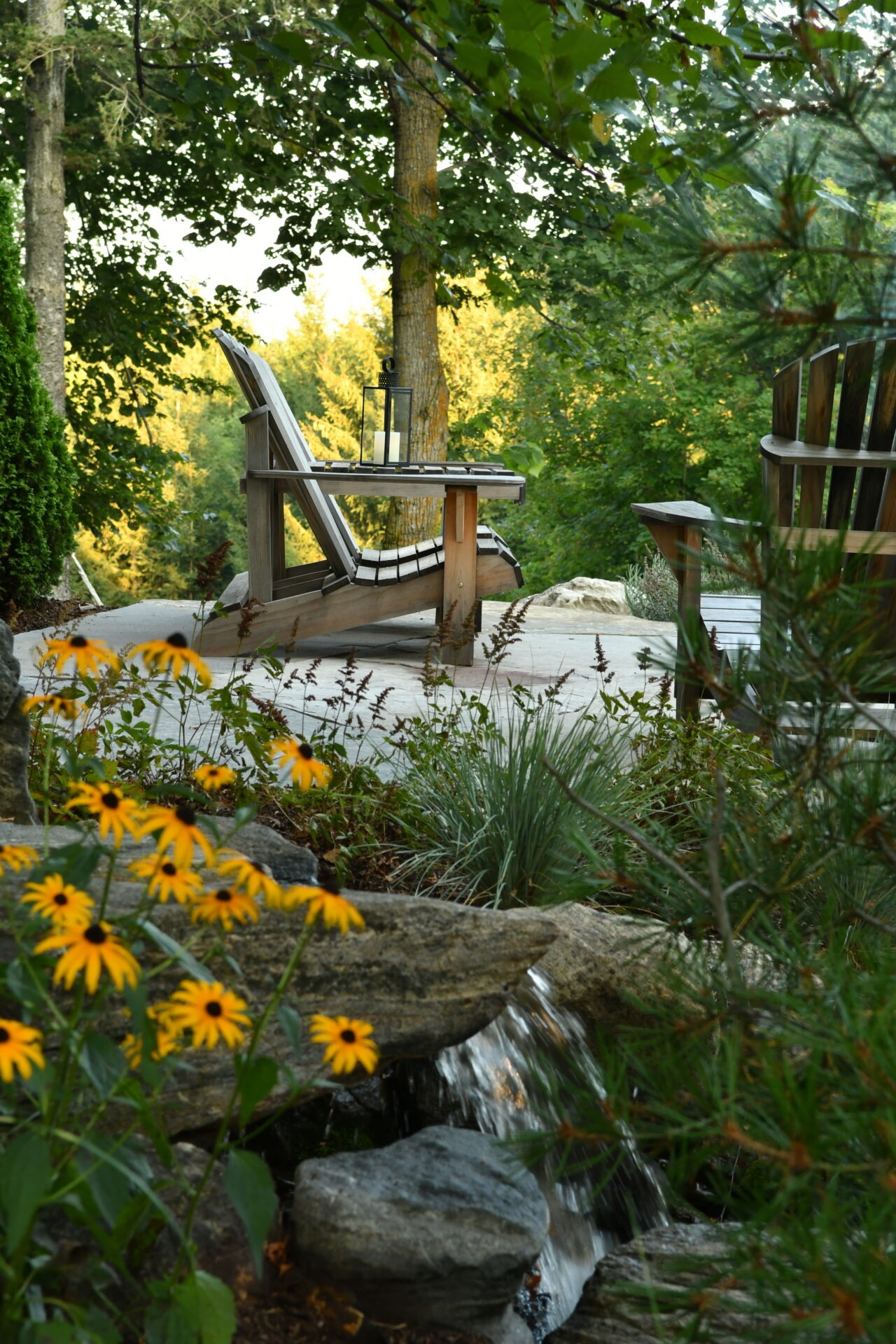 Wooden chairs on a patio overlook a serene forest, surrounded by lush greenery and yellow flowers beside a small waterfall feature.