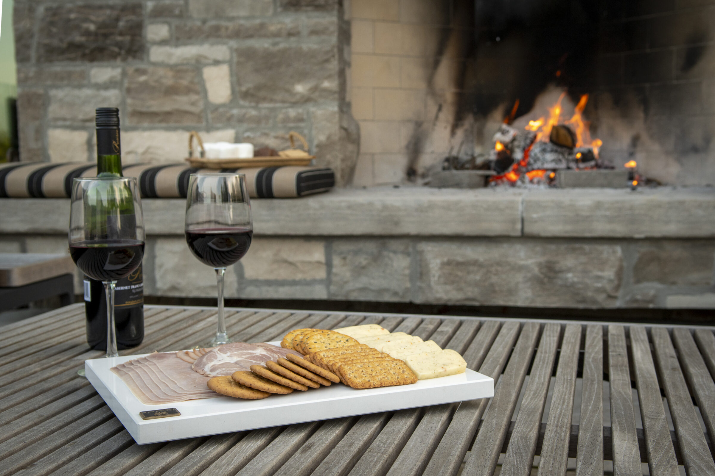 Two glasses of red wine, charcuterie, and crackers on a wooden table near a stone fireplace with visible flames and embers.