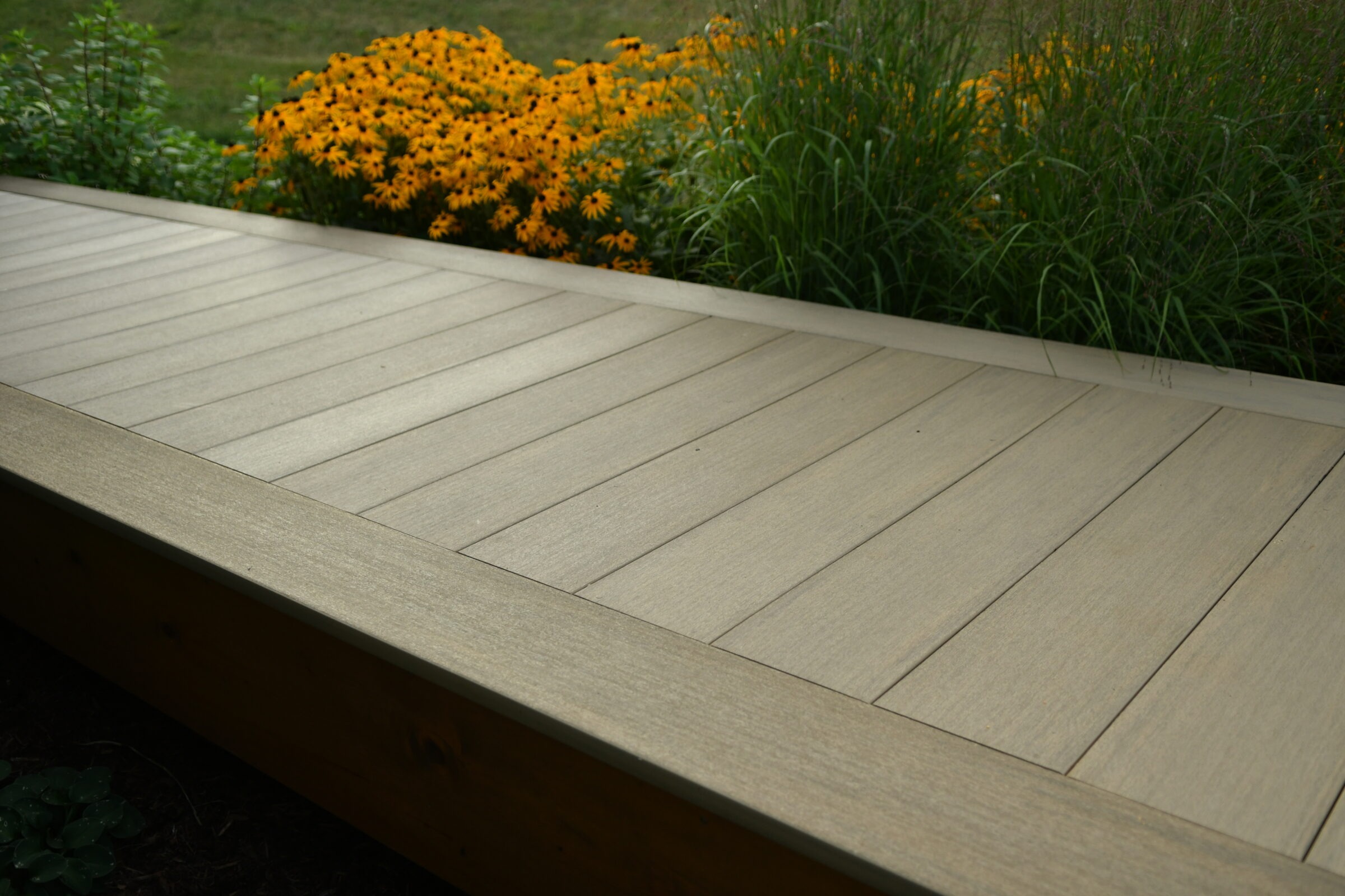 A wooden deck adjacent to vibrant yellow flowers and lush green plants, set against a backdrop of grassland landscape.
