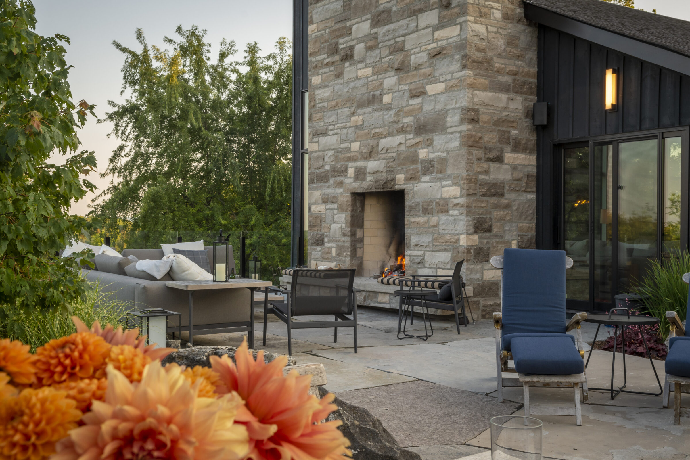 Patio with stone fireplace, blue lounge chairs, and blooming orange flowers. Comfortable seating areas surrounded by lush green trees, creating a serene atmosphere.