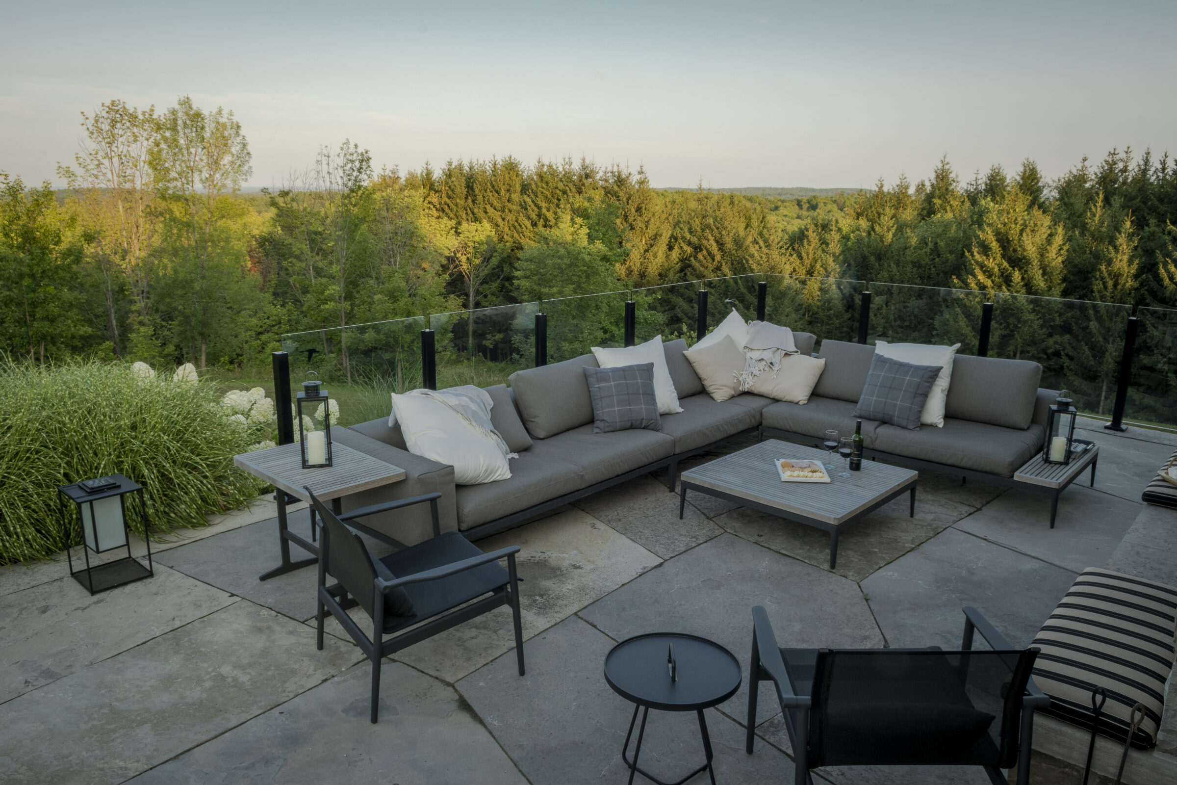 Spacious outdoor patio with grey sectional sofa, surrounded by lush greenery and trees. Glass railing provides a scenic woodland view.