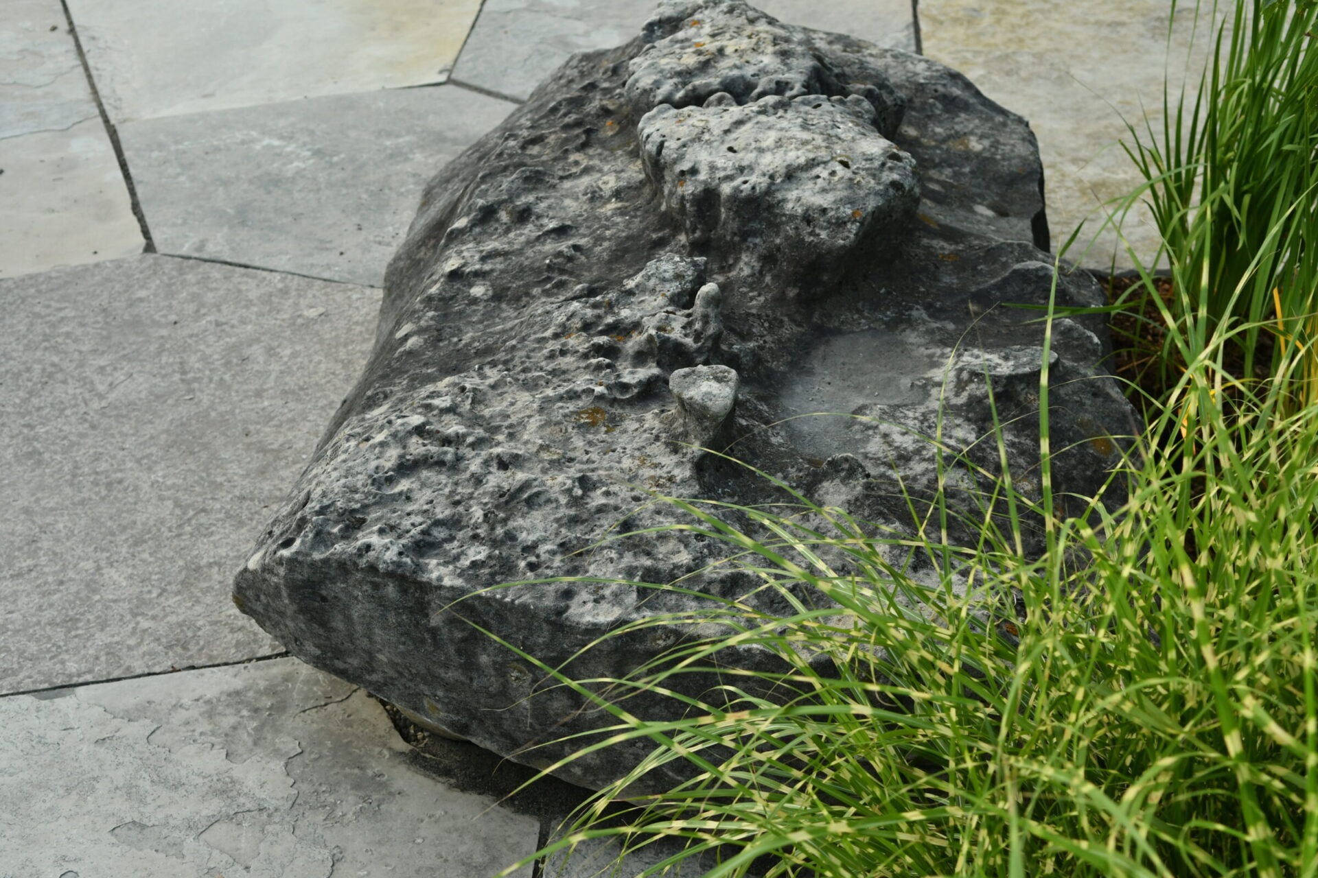 Rough-textured stone rests on a hexagonal tiled surface, surrounded by lush green grass, creating a natural, tranquil scene.