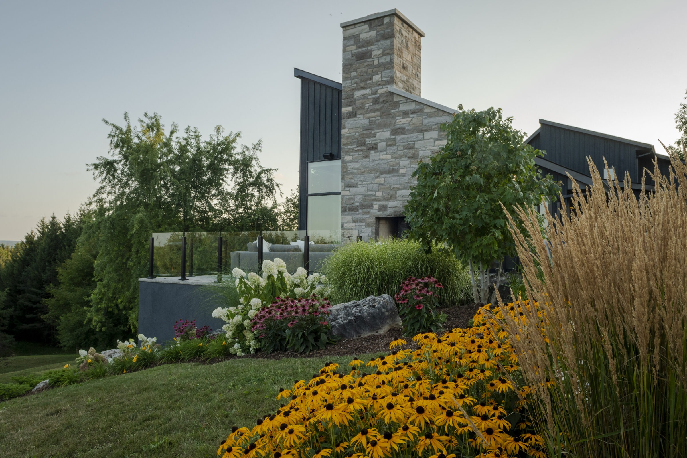 Contemporary house with stone chimney, surrounded by vibrant garden featuring yellow and white flowers, amidst lush green trees and manicured lawn.