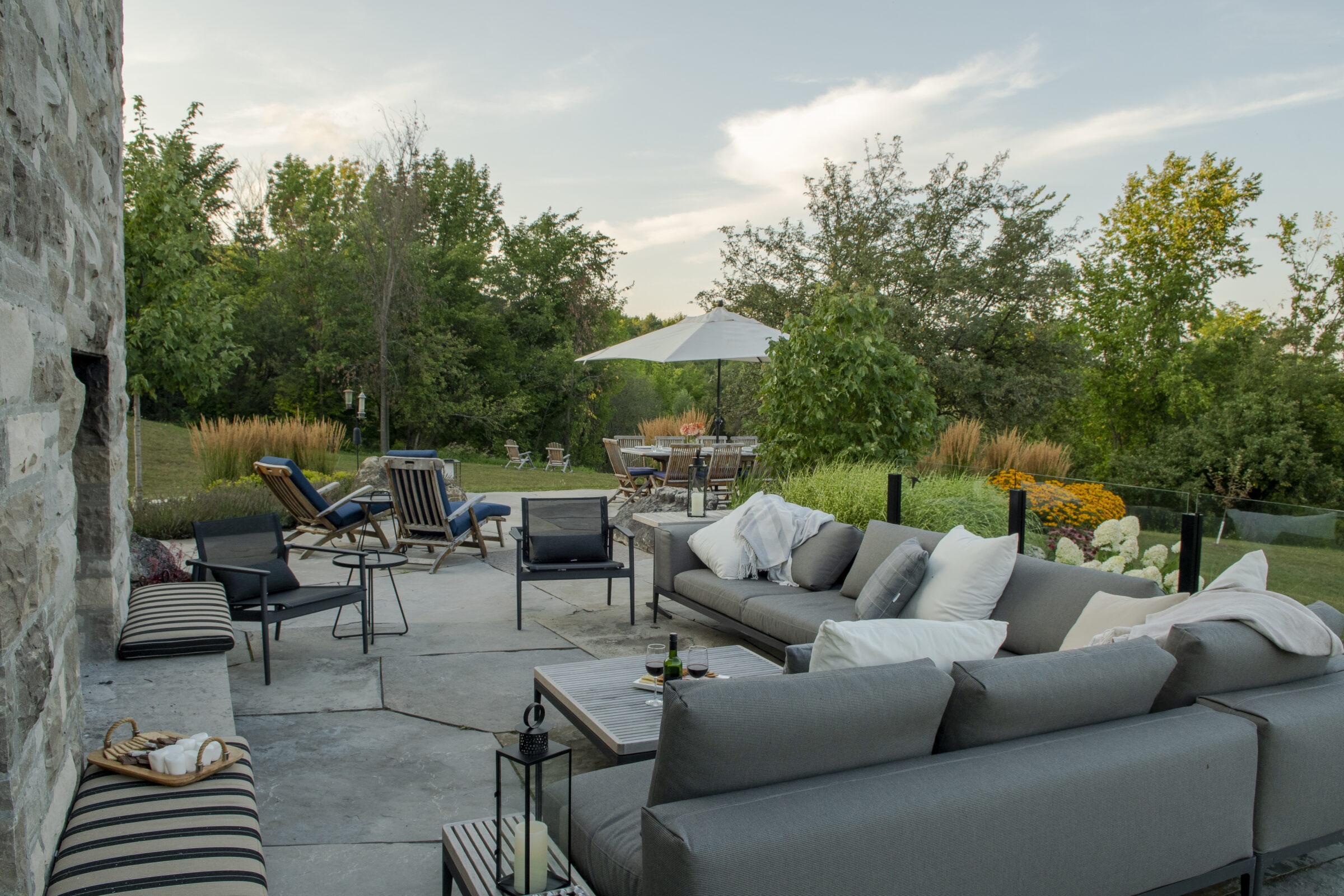 A cozy patio scene with grey sofas, a large umbrella, stone walls, and lush greenery, creating a serene outdoor relaxation space.