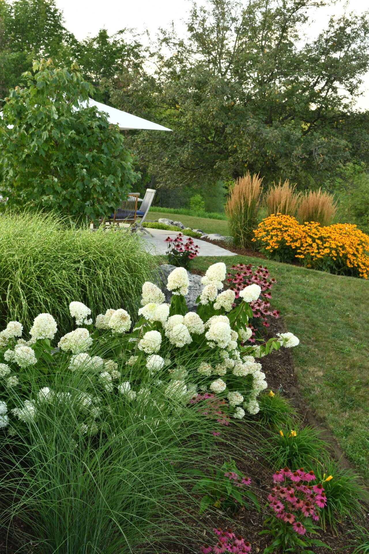 Lush garden with colorful flowers, hydrangeas, and decorative grasses. A patio area features outdoor seating under a canopy, surrounded by greenery.