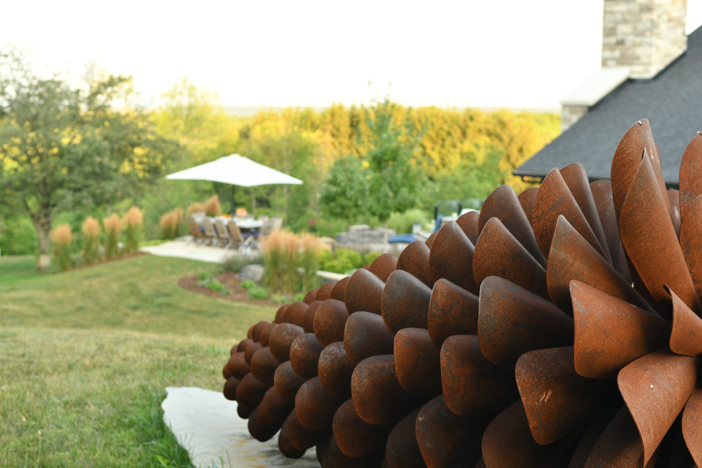 Outdoor scene with large rusted sculpture in foreground, patio with table and chairs under umbrella, surrounded by greenery and garden landscape.