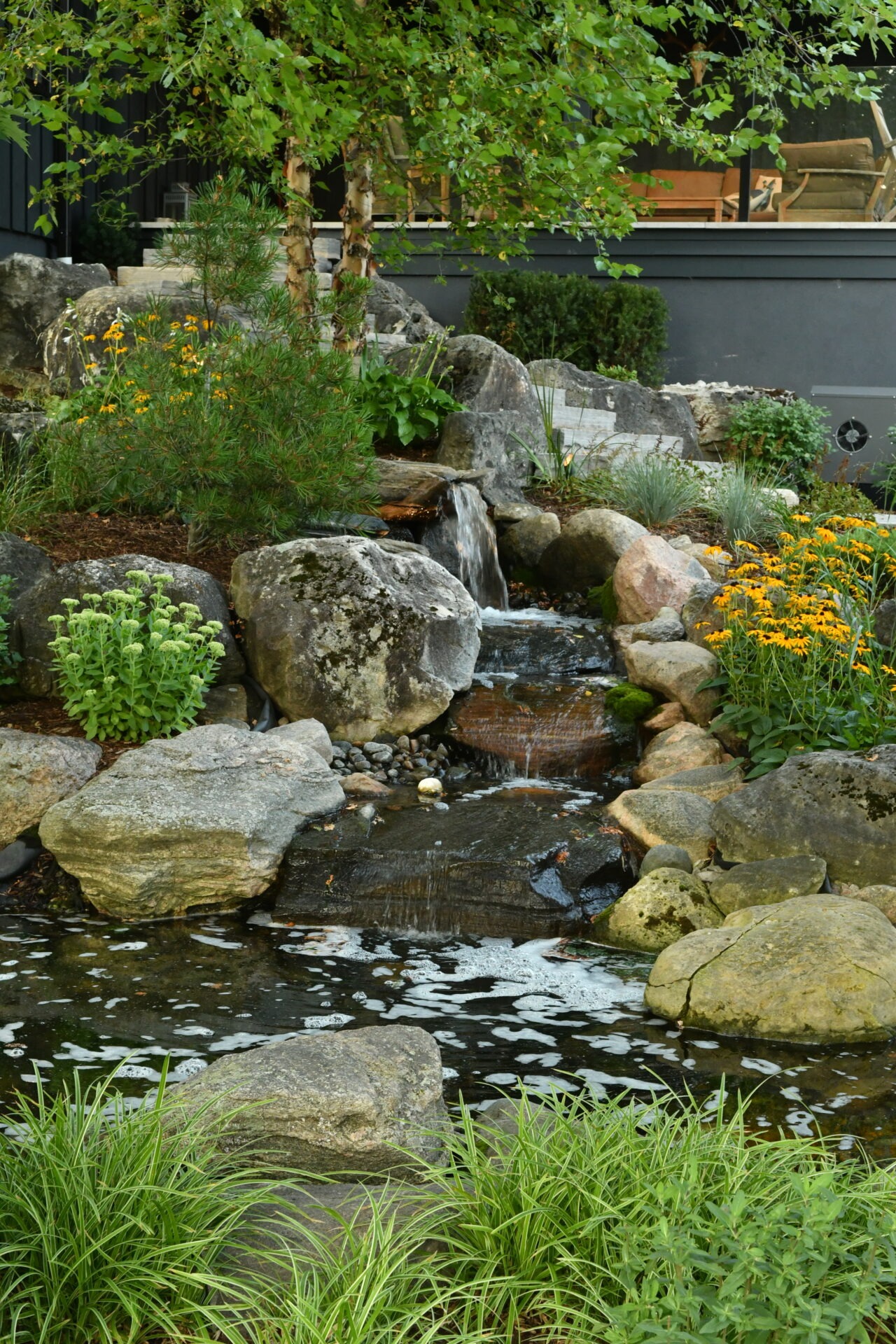 A small waterfall flows over rocks into a pond surrounded by greenery and flowers, with a building visible in the background.