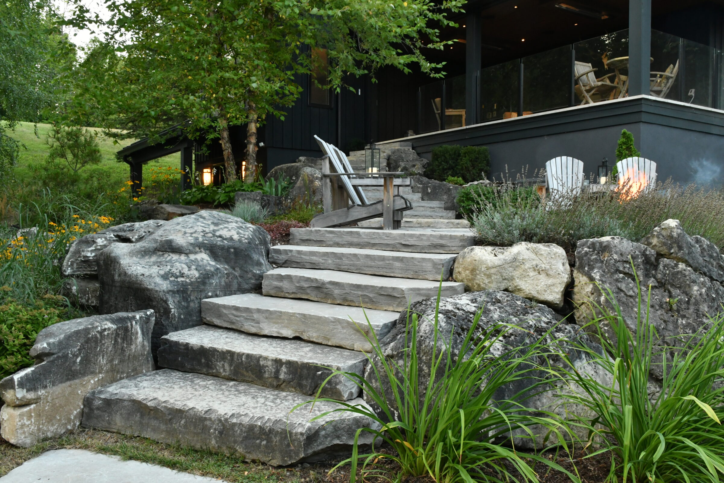 Stone steps surrounded by rocks and lush greenery lead to a cozy patio with chairs and a fire pit, nestled in nature.