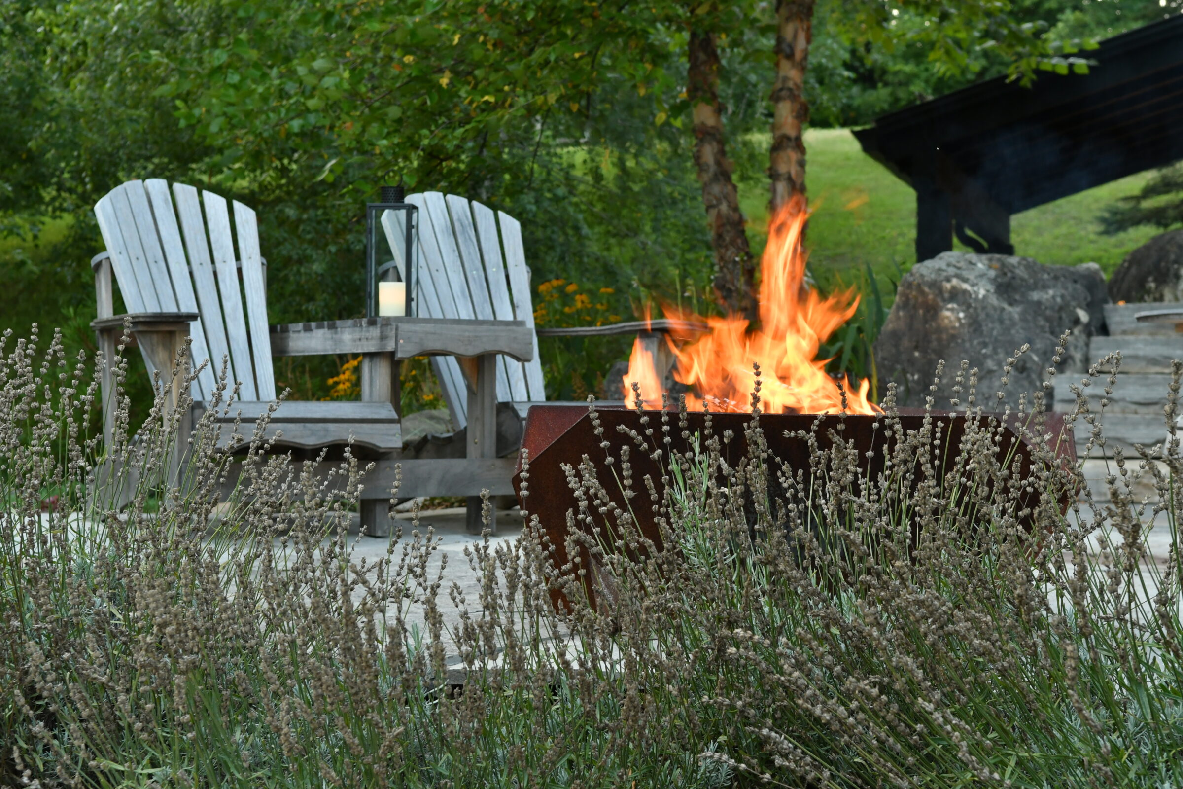 Two wooden chairs by a lit fire pit in a garden setting, surrounded by greenery and lavender plants, with a lantern nearby.